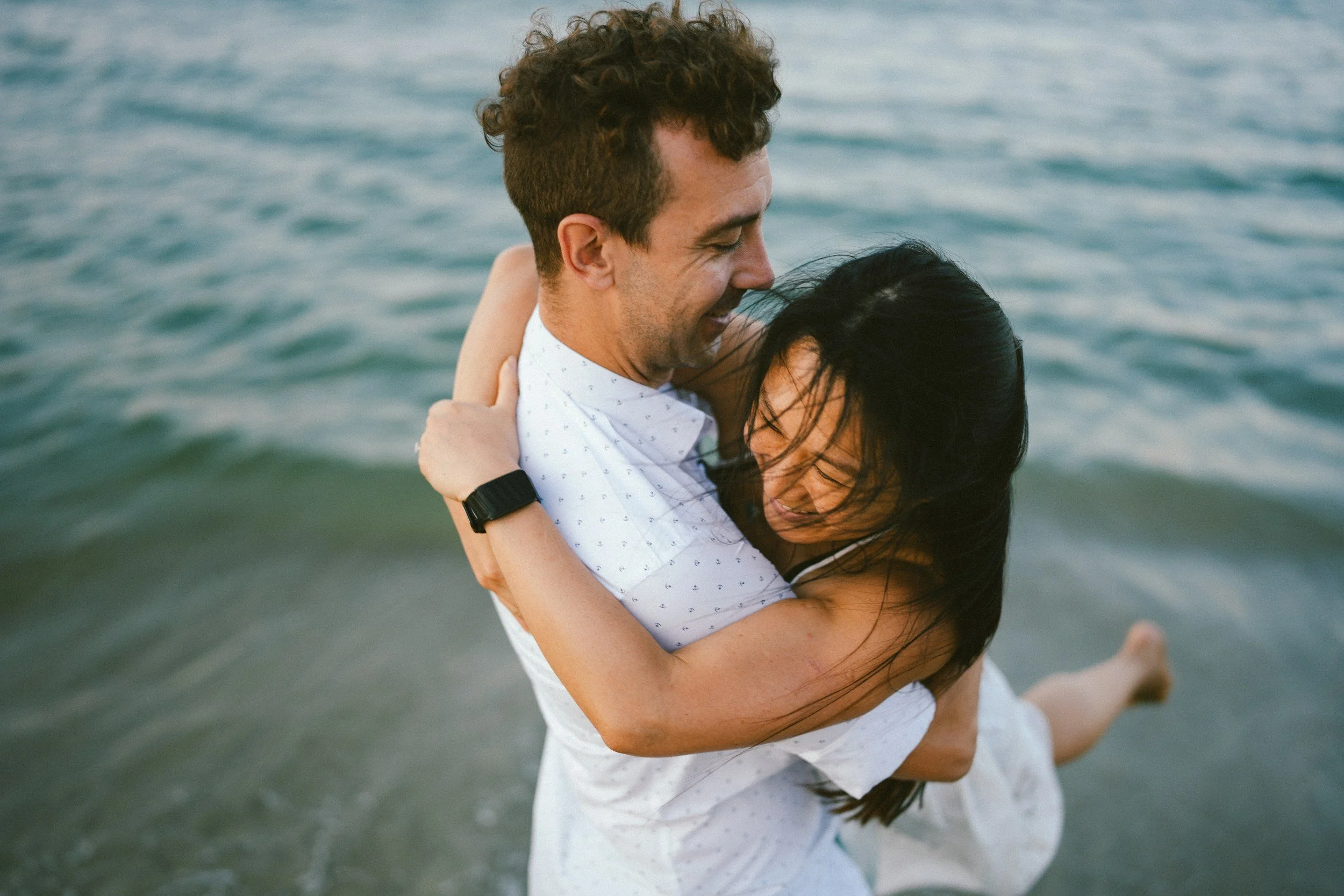 A man and woman embracing in shallow ocean water, smiling, with the woman wrapping her arms around the man's neck.
