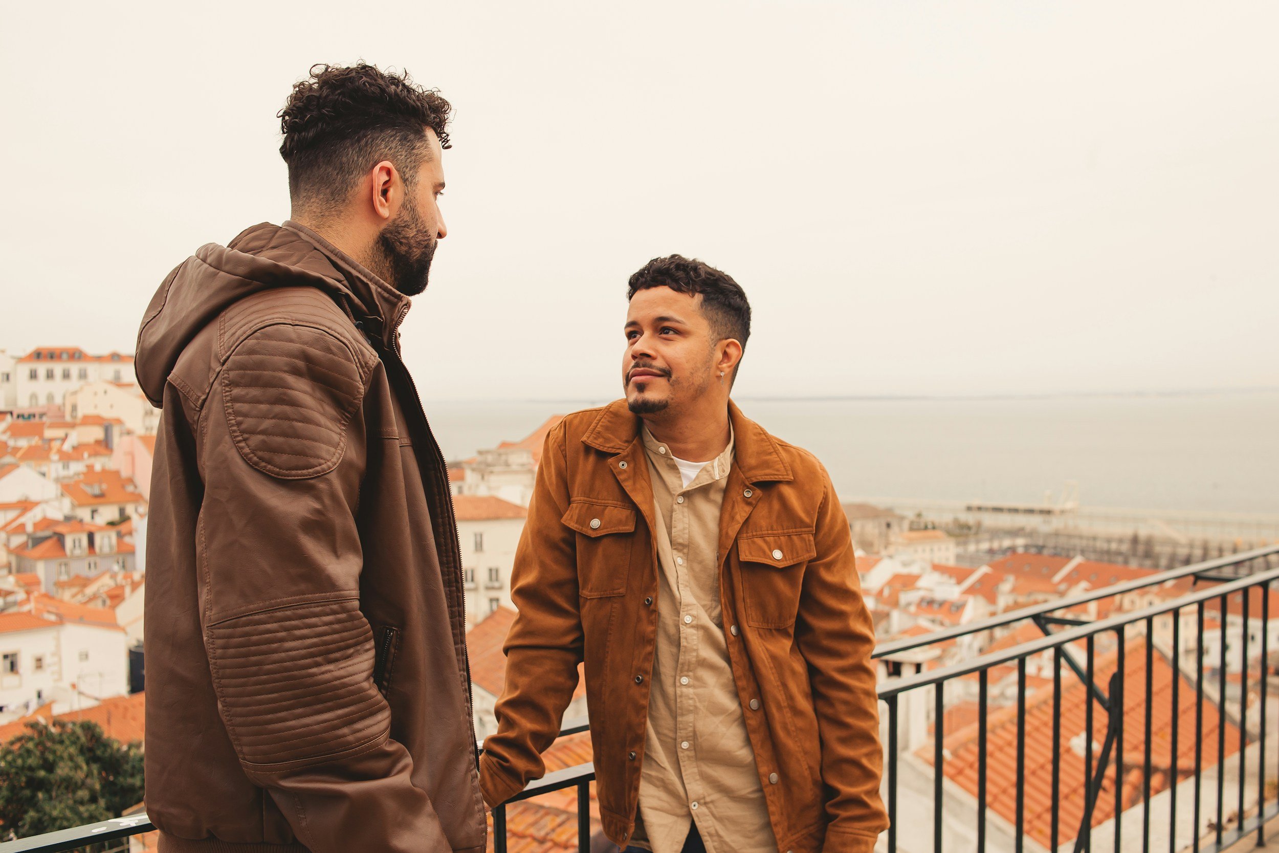 Two men standing on a balcony overlooking a city with orange rooftops, engaged in a conversation, with a body of water in the background under a cloudy sky.