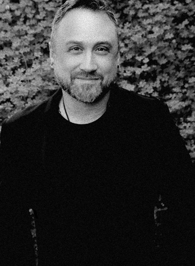 A middle-aged man with light hair and a beard smiling, wearing a black shirt, outdoors with blurred foliage in the background.