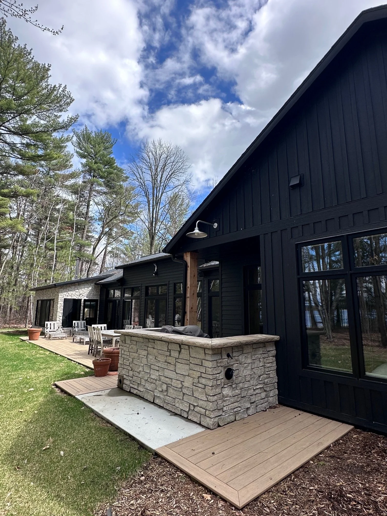 Backyard patio with outdoor dining furniture, potted plants, a built-in stone grill, and a black wooden house with large windows, surrounded by trees and clear sky.