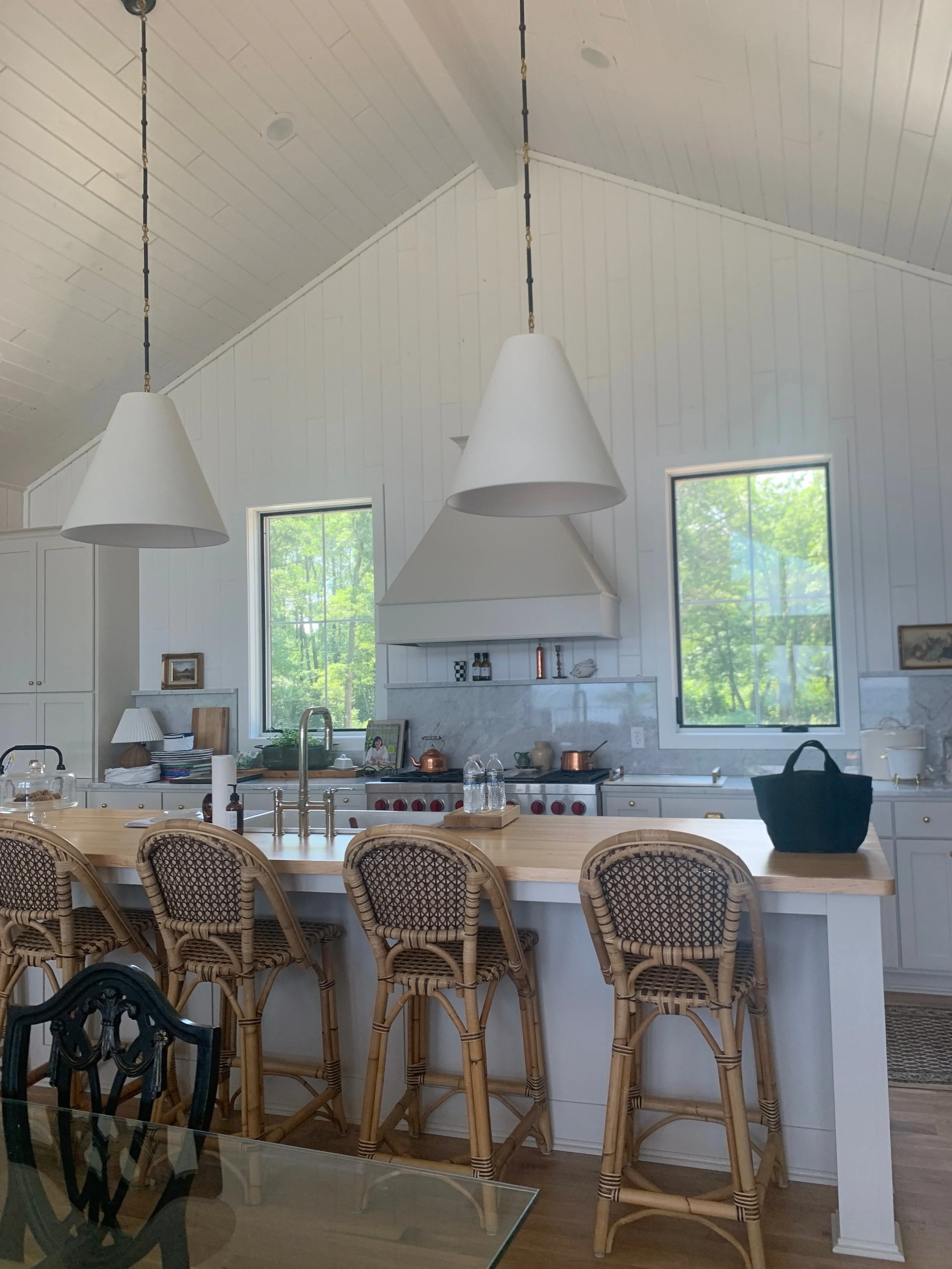 Bright kitchen with white cabinets, two large windows, and a light wood island with four wicker bar stools. Pendant lights hang from the vaulted ceiling, and there are various kitchen items on the counter and shelves.