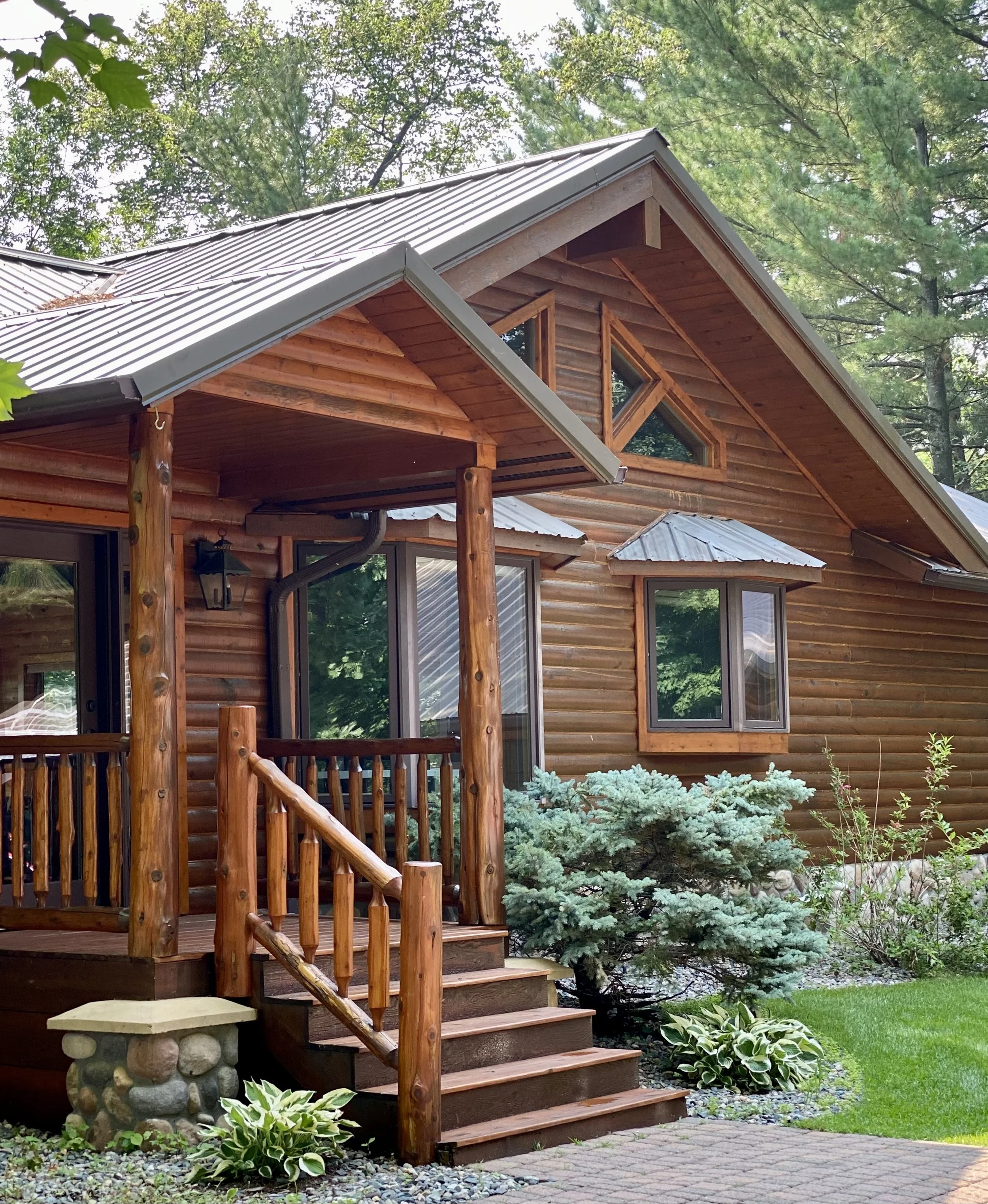 A wooden house with a porch, surrounded by trees and a garden, featuring a stone foundation, metal roof, and large windows.