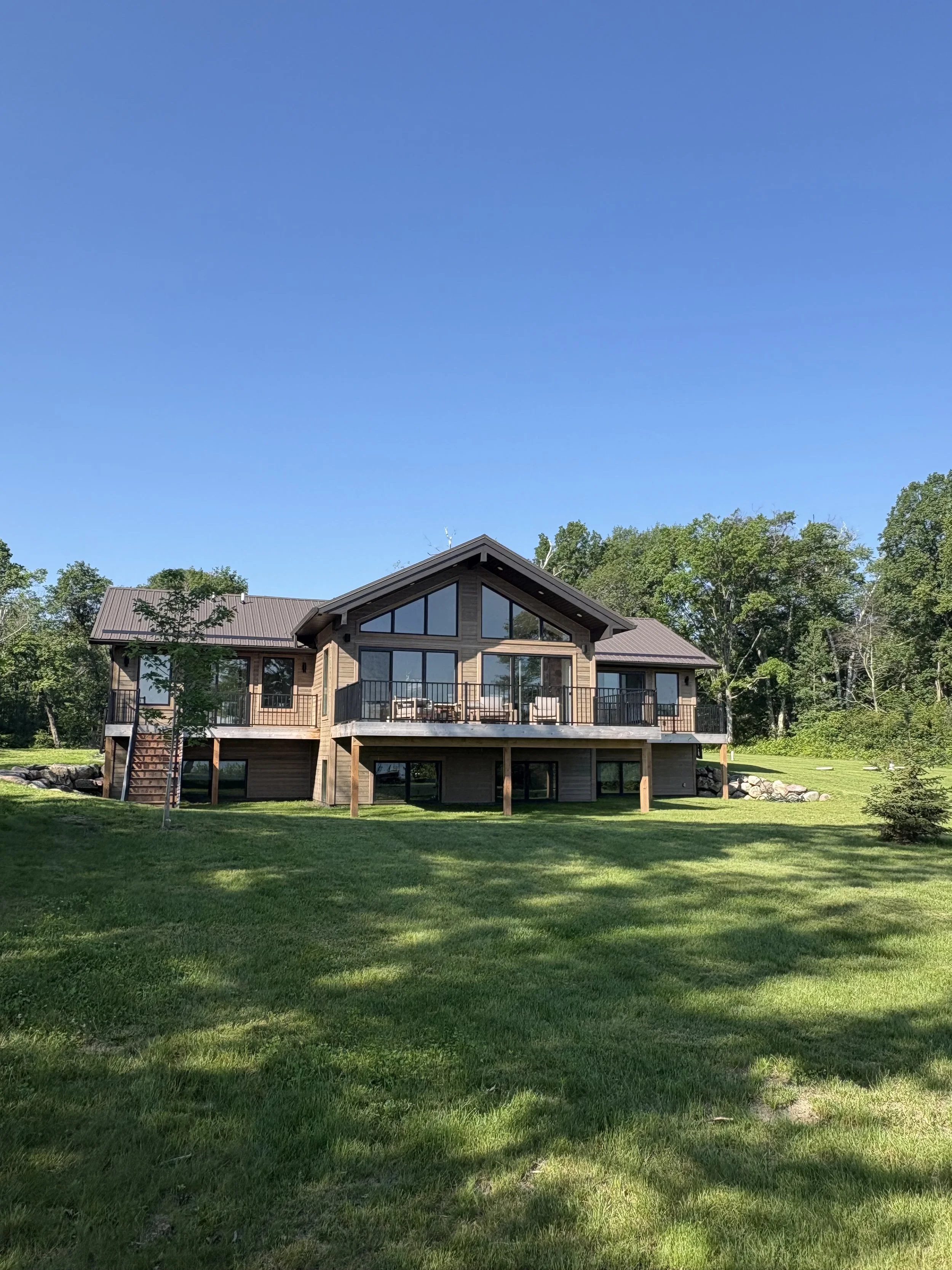 Modern two-story house with large glass windows and a spacious deck on the back, surrounded by green lawn and trees under a clear blue sky.