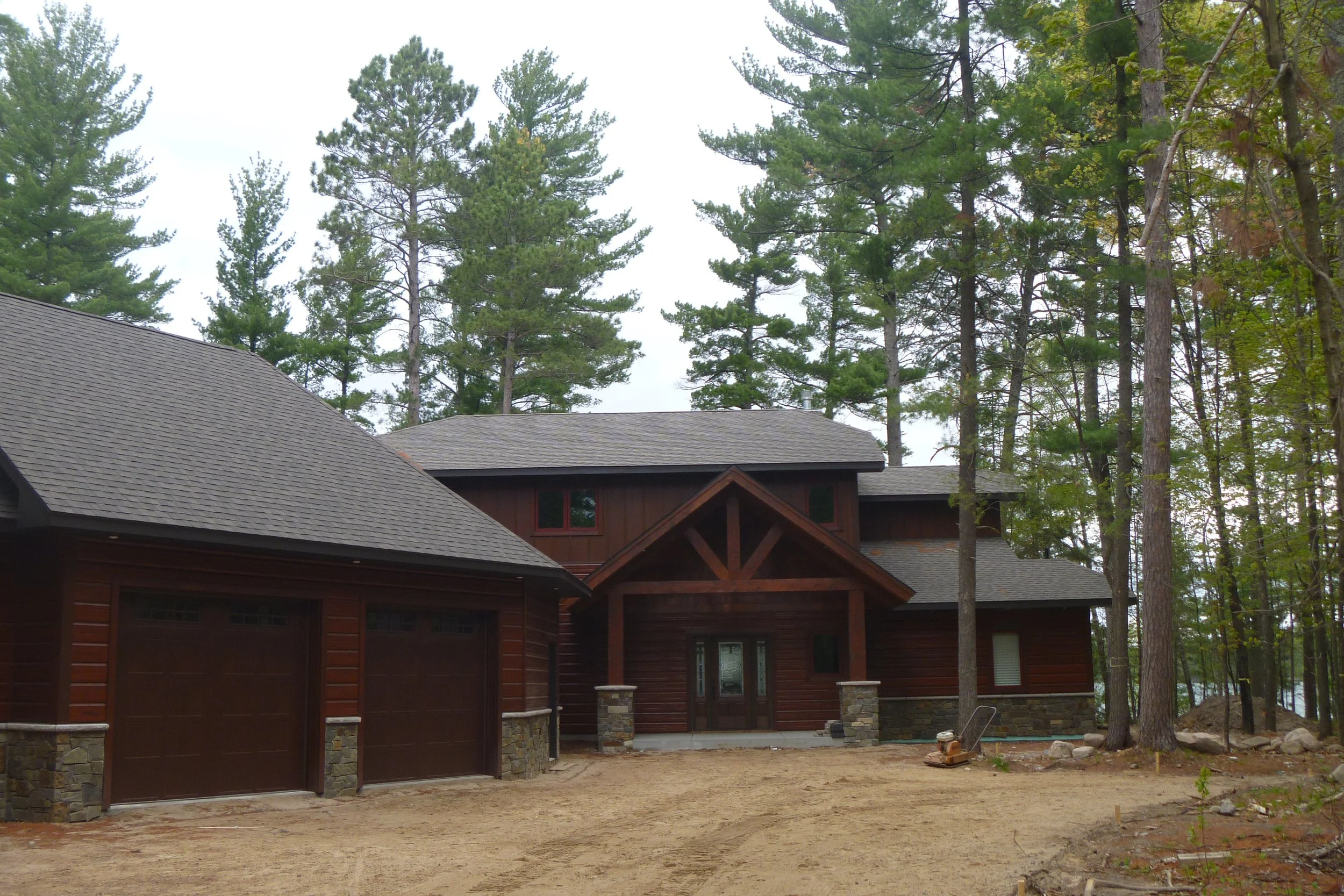 A house under construction in a wooded area, with a newly paved driveway, in a forested setting with tall trees surrounding the house.
