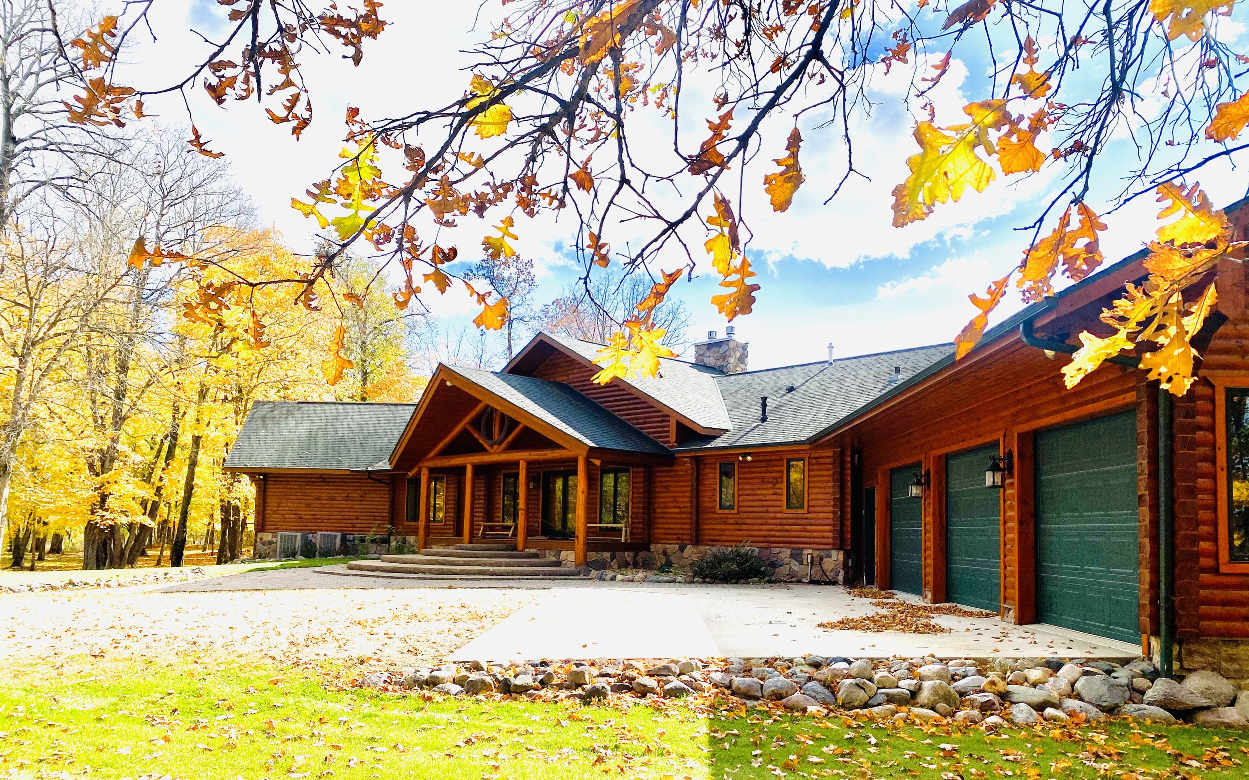 A wooden house with a green garage door, stone accents, and a porch with steps, surrounded by trees with autumn leaves.