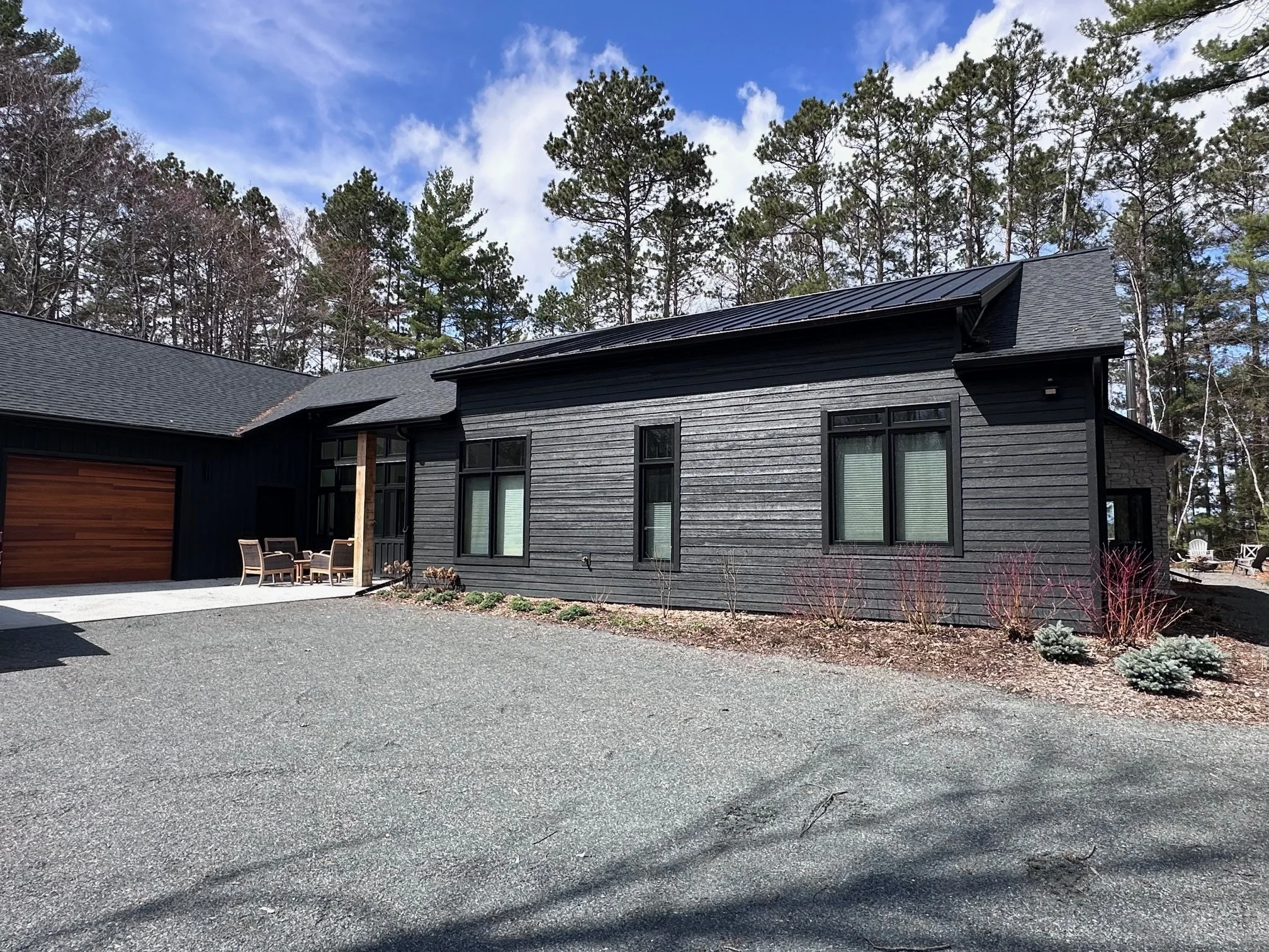 A modern black wooden house with large windows and a grey shingled roof. There is a gravel driveway and a small outdoor sitting area with chairs. The house is surrounded by trees and a partly cloudy sky.