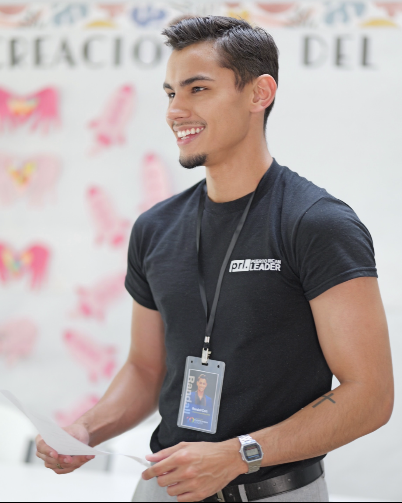 A young man with dark hair and a goatee smiling, wearing a black t-shirt with a logo and a lanyard, standing in front of a white background with colorful butterflies and text, holding a piece of paper.
