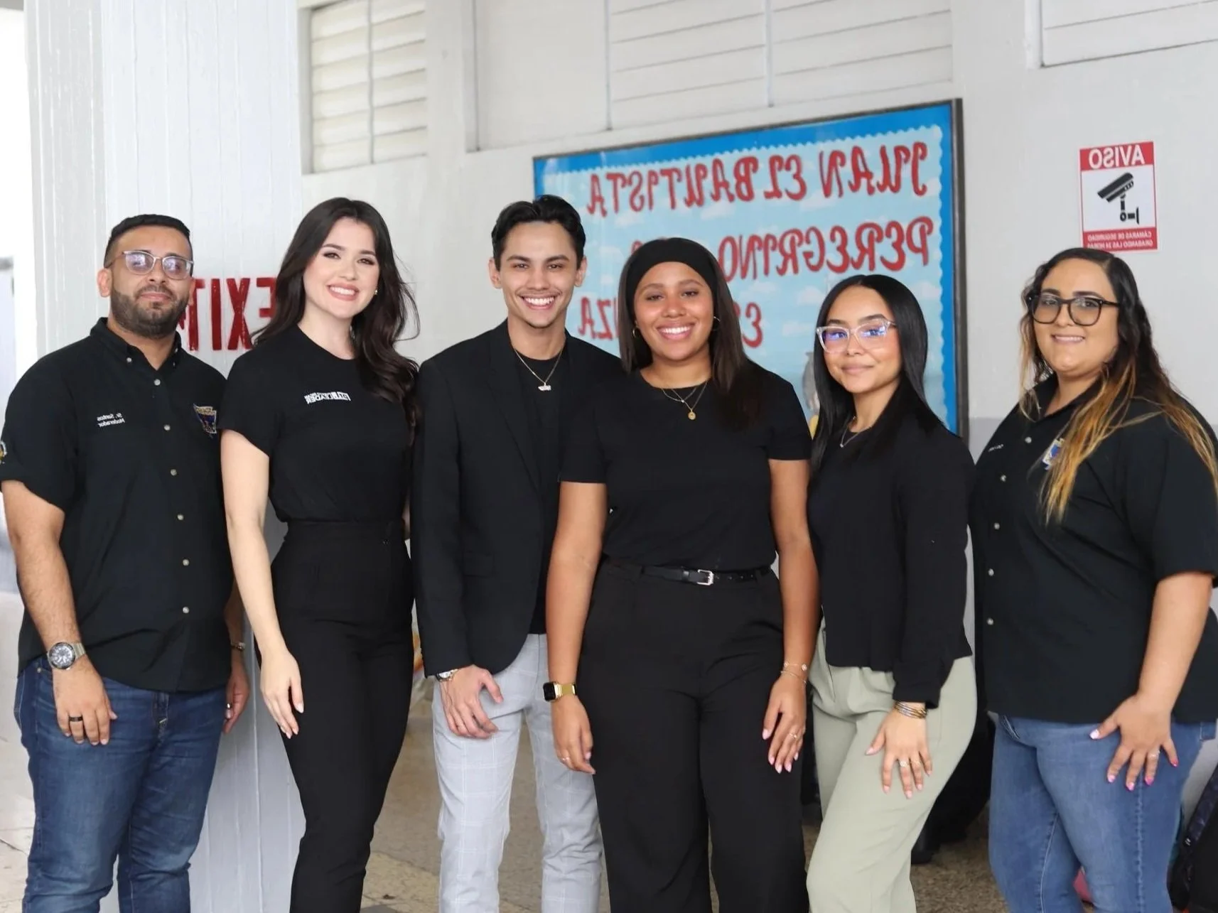 Group of six diverse young adults standing together indoors, smiling at the camera. They are dressed in black and light-colored clothing, with a blue bulletin board and a red fire alarm sign in the background.