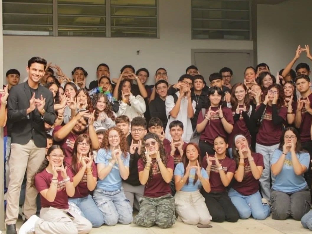 Group of students and a teacher gathered for a photo in a school setting, some making hand signs and smiling.