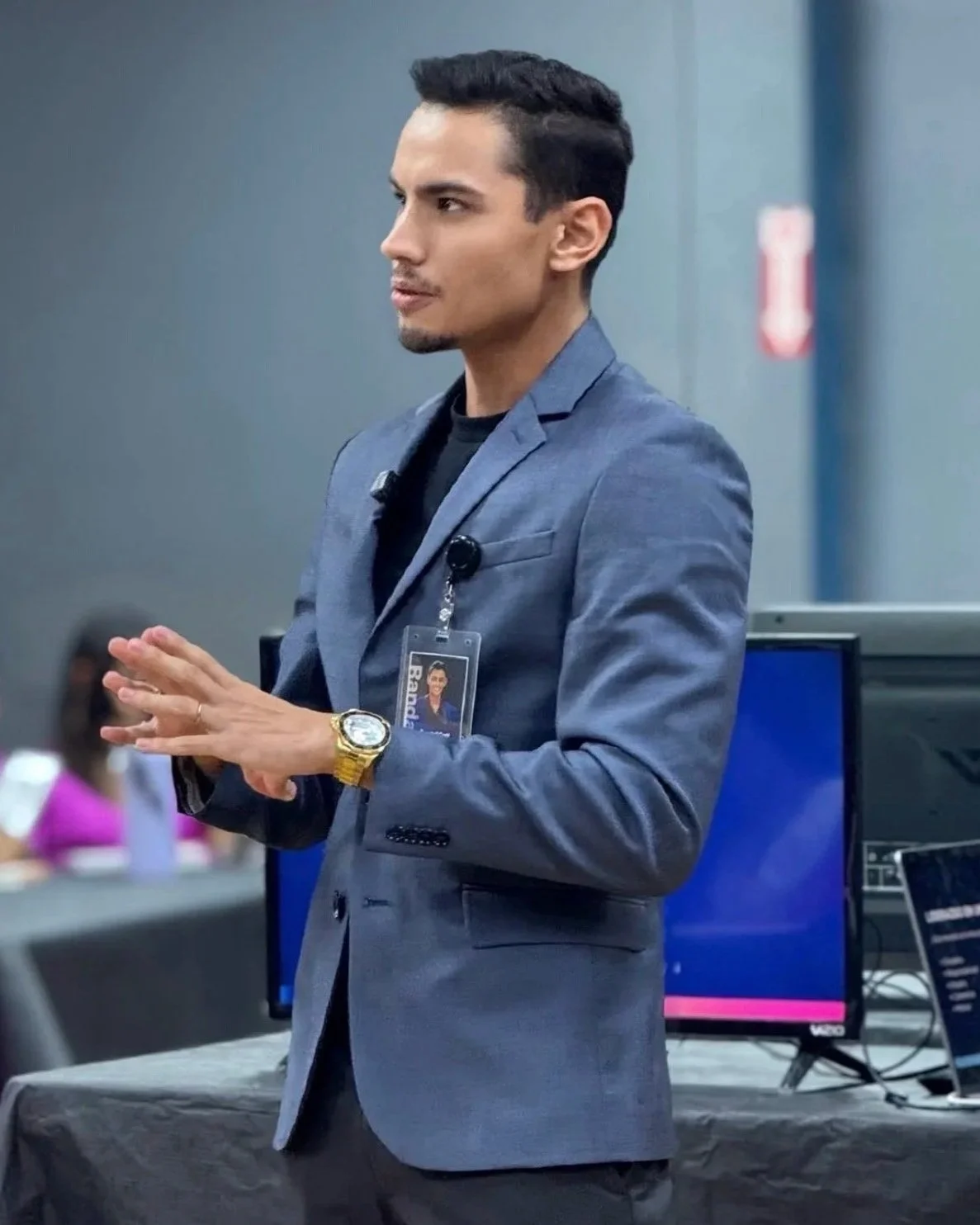 A man in a gray suit with a badge, gold watch, and bracelet, speaking in front of an audience at a conference or presentation.