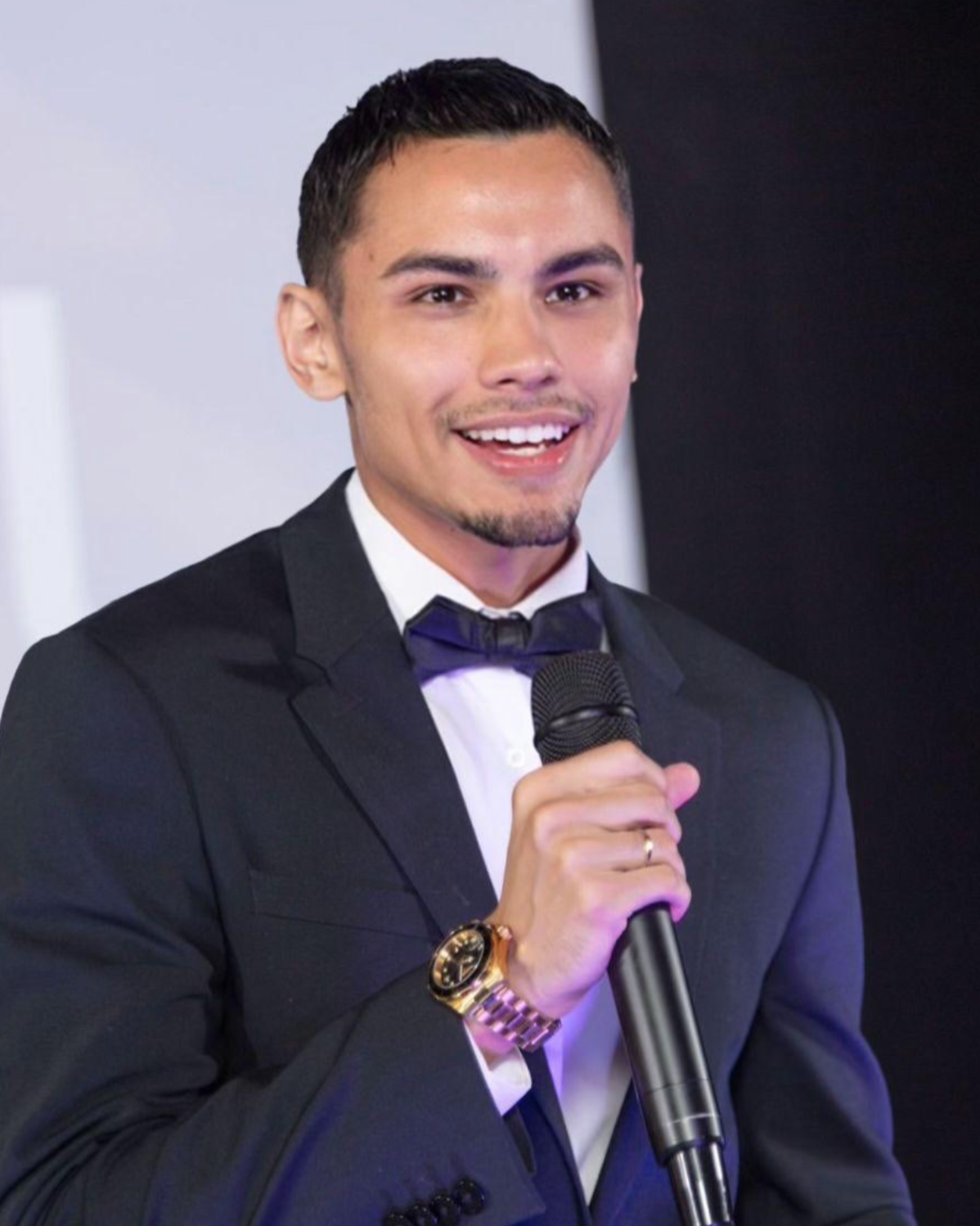 A man wearing a black tuxedo and bow tie, holding a microphone, smiling, in an indoor setting.