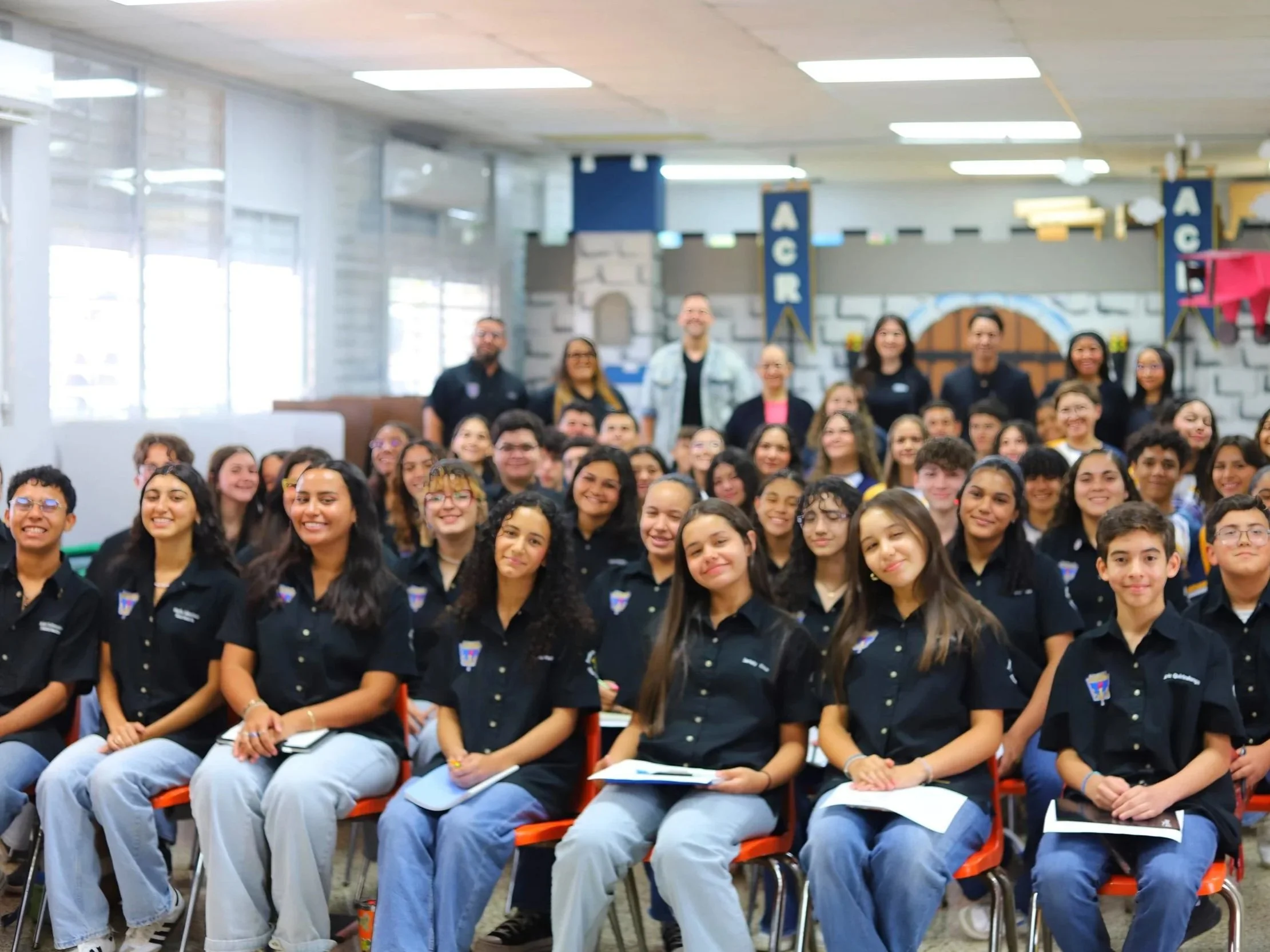 A large group of students and teachers gathered in a school auditorium, sitting on chairs and smiling for the photo, with some standing in the background and banners hanging on the walls.