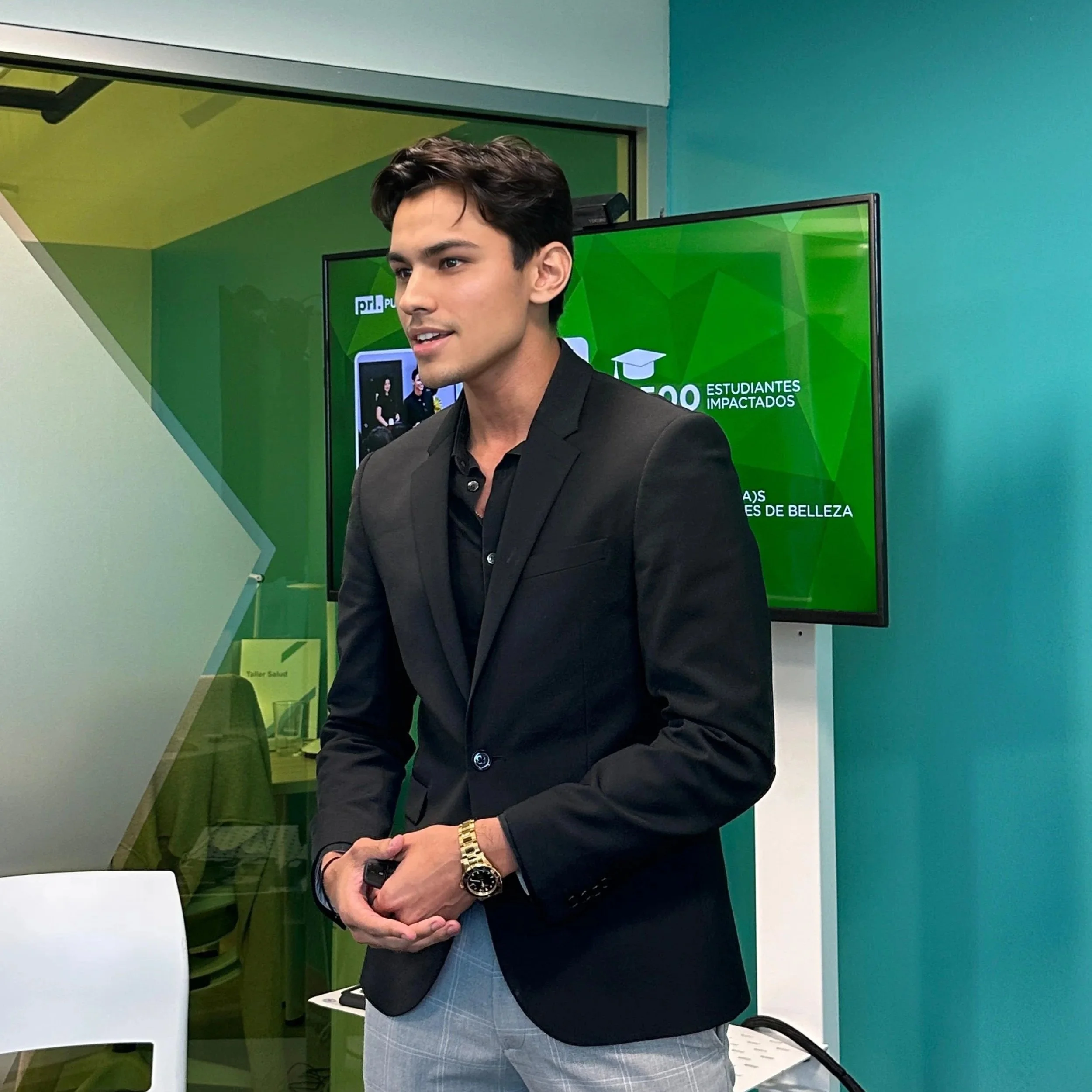 A young man dressed in a black blazer and checked trousers is standing in front of a green and blue wall with a screen, during a presentation or seminar. He has dark, styled hair, and is wearing a gold watch on his left wrist.