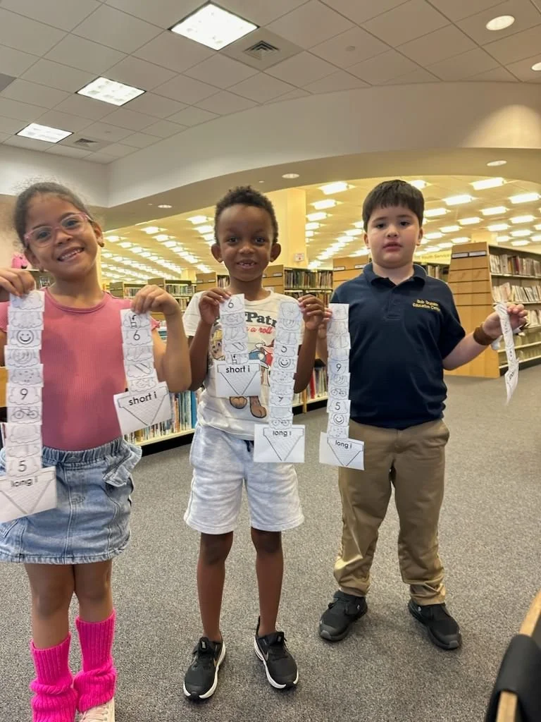 Three children standing in a library, holding paper chains with numbers and shapes, smiling and showing their crafts.