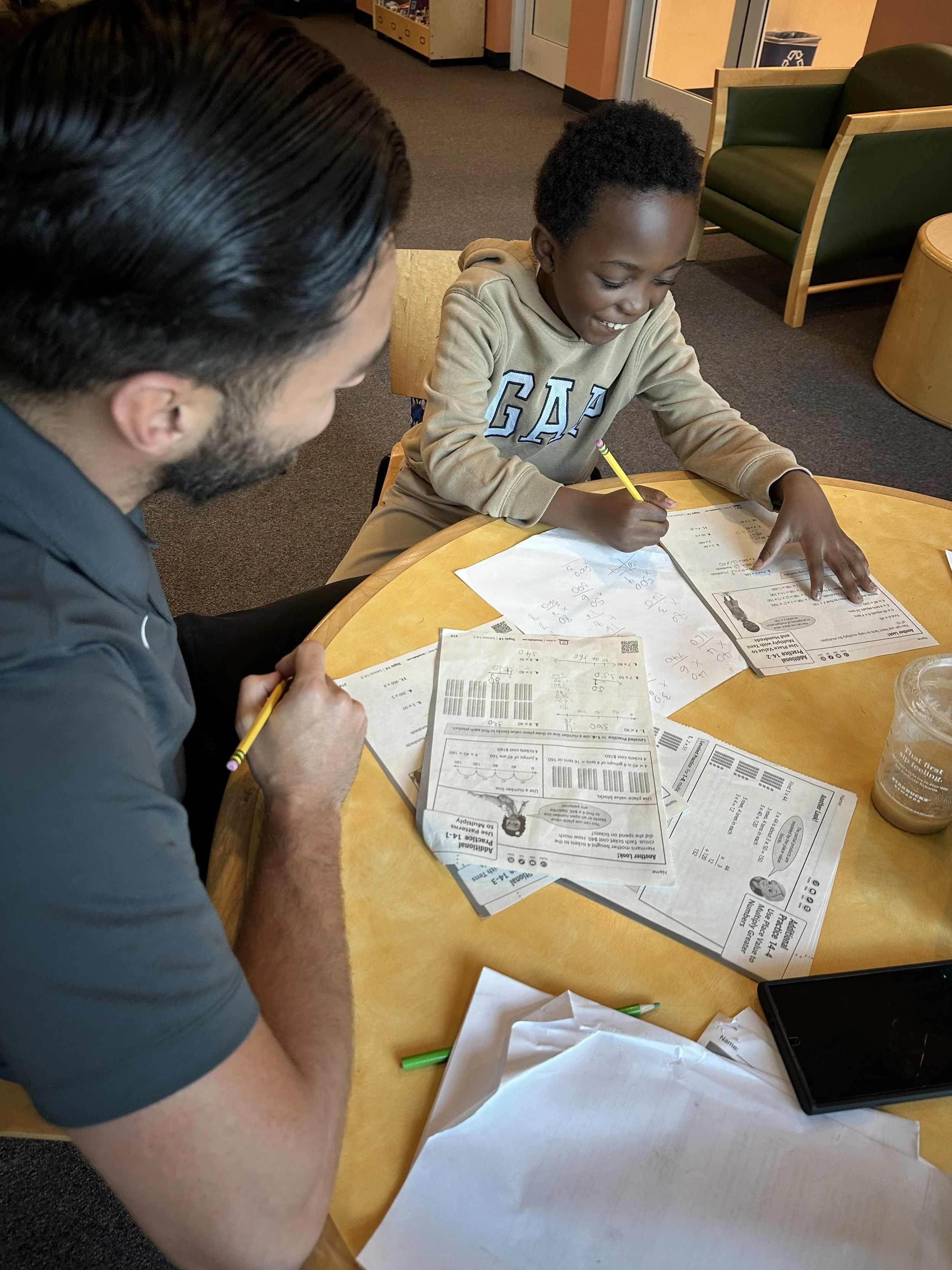 A young boy sitting at a round table with an adult man, working on papers with math problems and barcodes, while the man looks on. There are drinks, a phone, and various papers on the table.