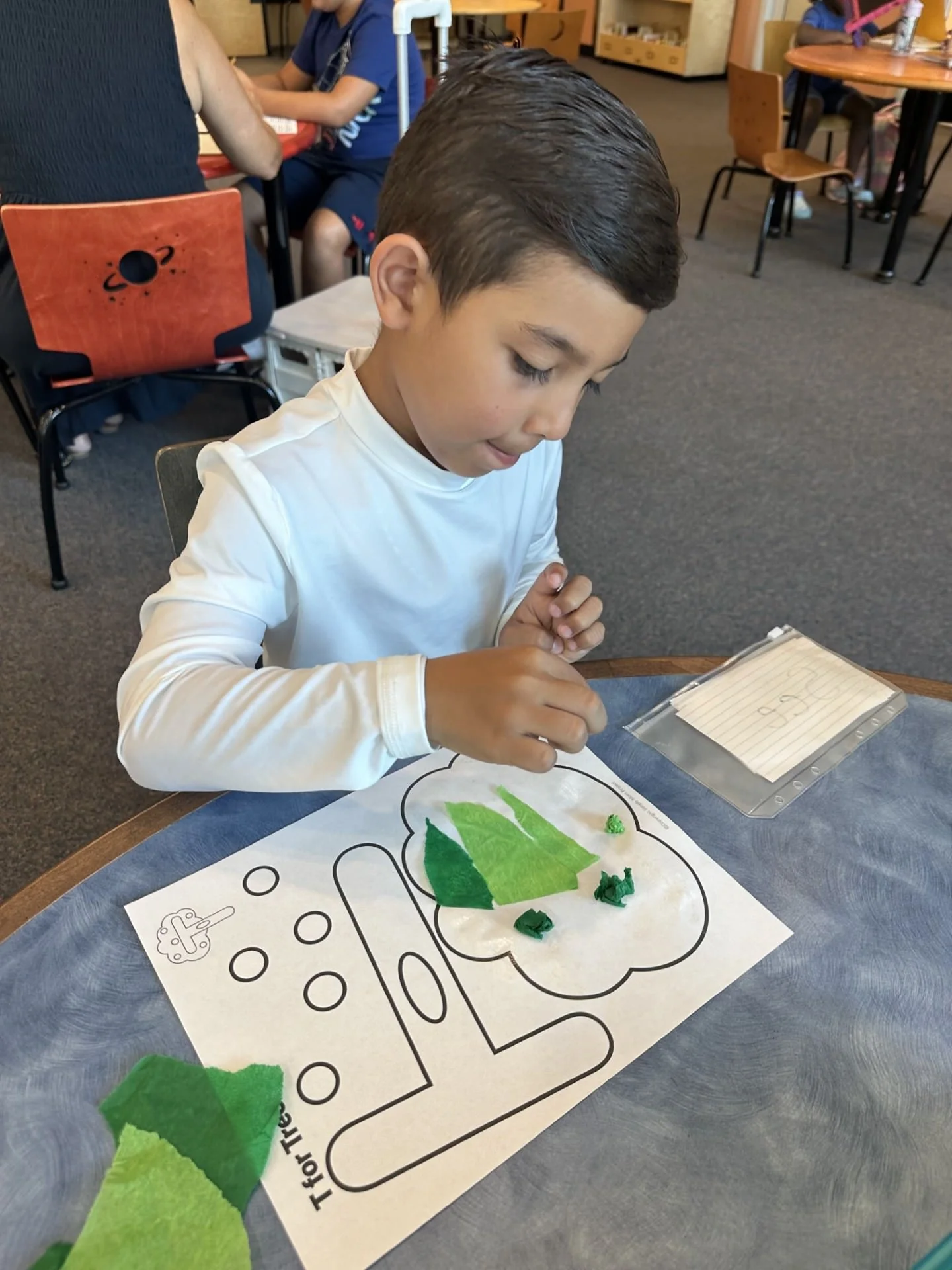 A young boy with dark hair and a white long-sleeve shirt is decorating a large printed coloring sheet with green tissue paper pieces, sitting at a table in a classroom or activity room.