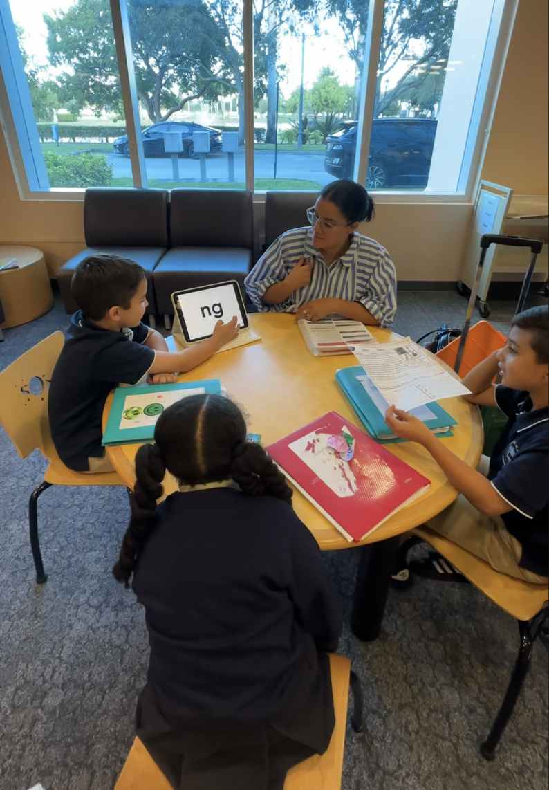 A teacher and four children seated around a yellow table in a classroom. The teacher is holding paperwork and looking at one of the children, who is holding a tablet with the letters 'ng' on it. The children have open notebooks and folders on the table, and a large window shows cars and trees outside.