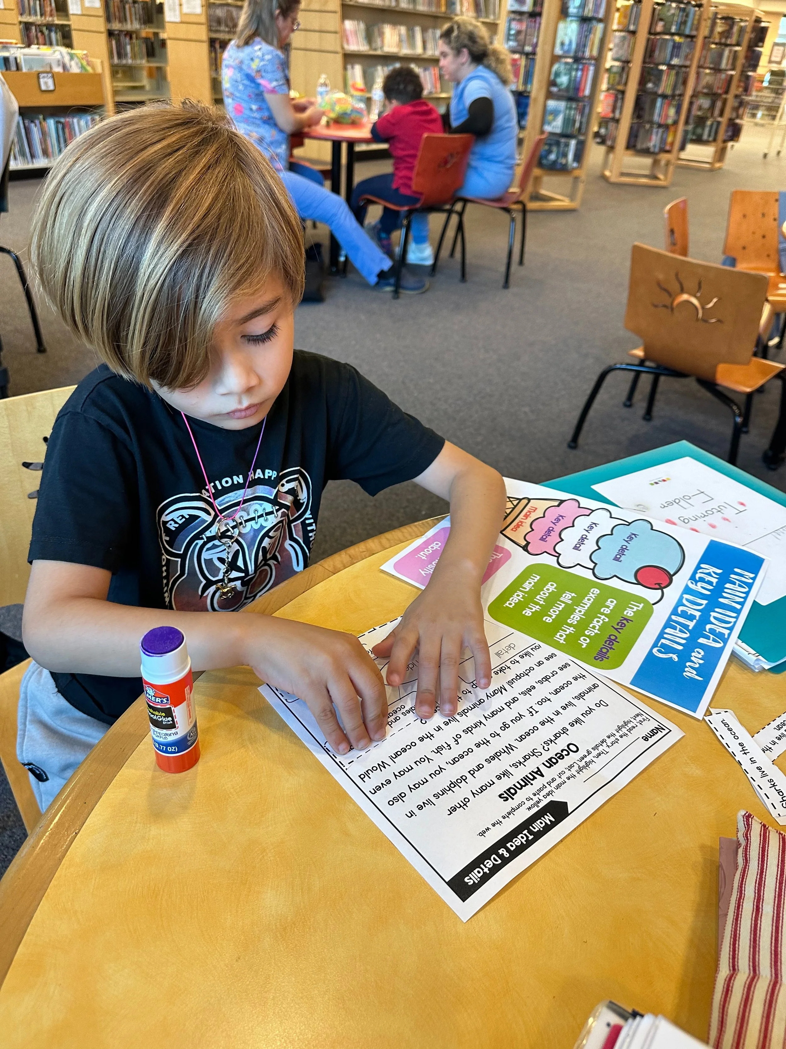 A young boy working on a reading worksheet at a library table, with classroom and bookshelves in the background.
