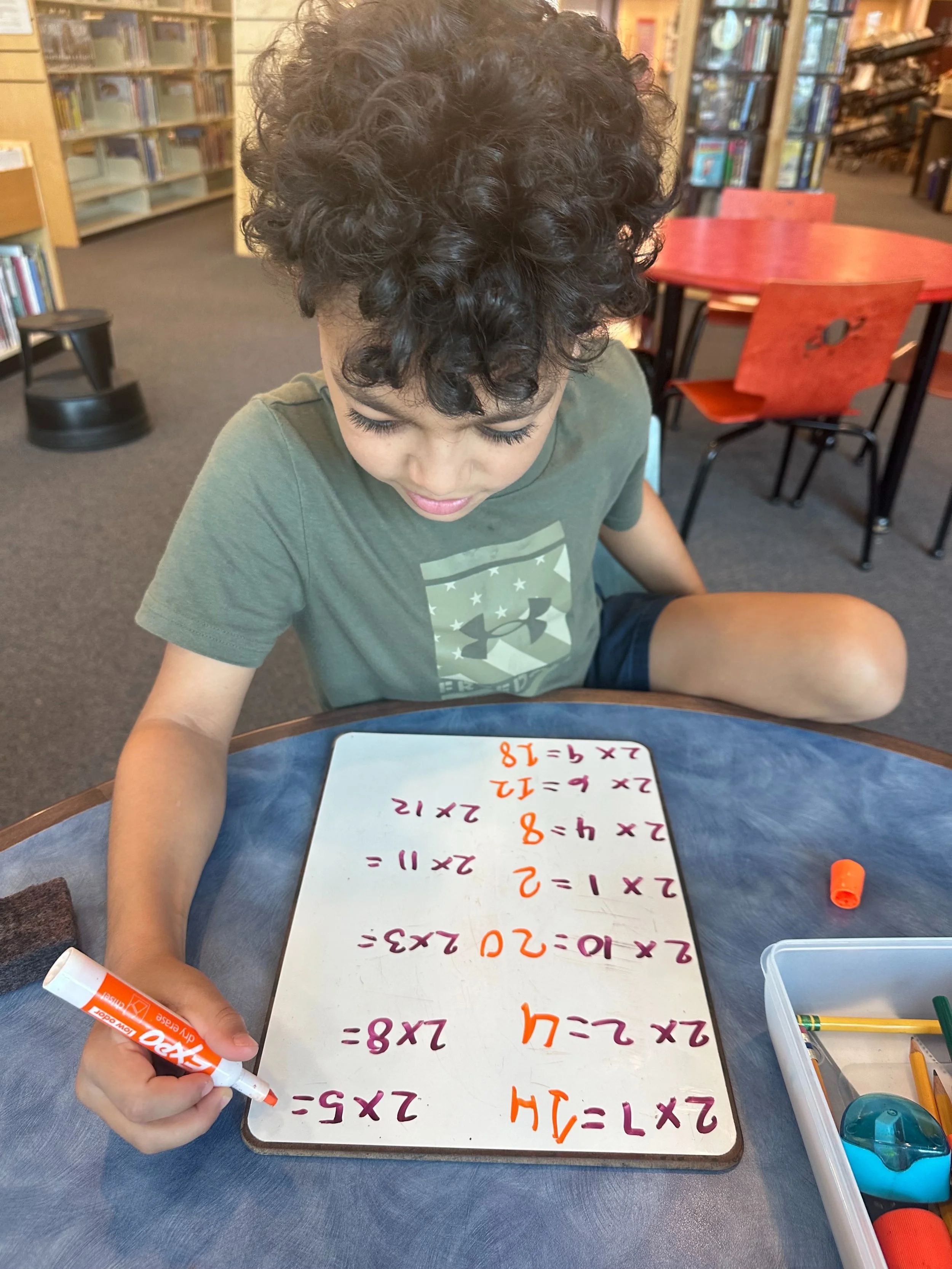 A young boy sitting at a table in a library, working on math problems with a whiteboard and markers.