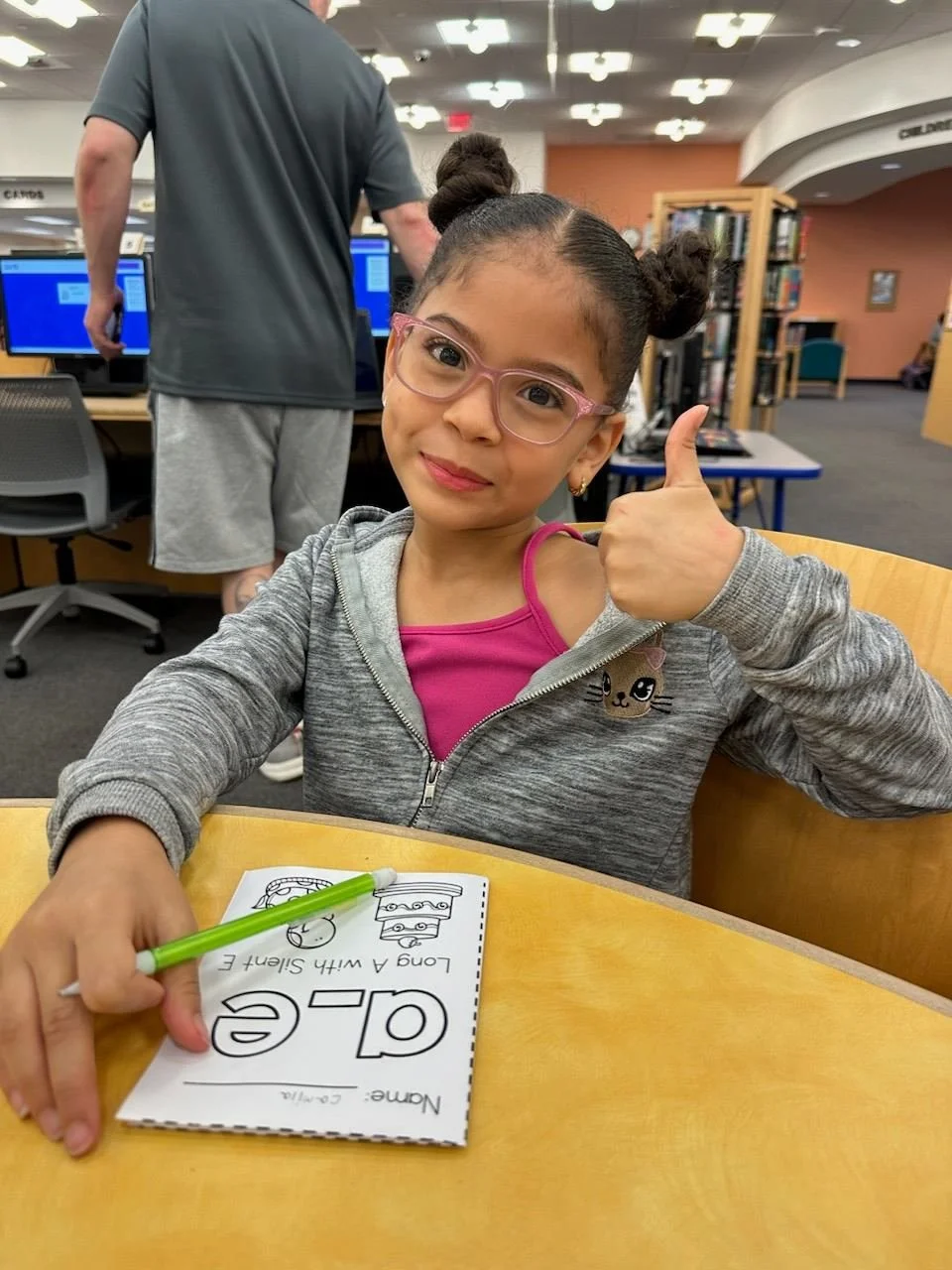 A young girl with glasses giving a thumbs up, sitting at a table with a coloring book and a green marker inside a library.