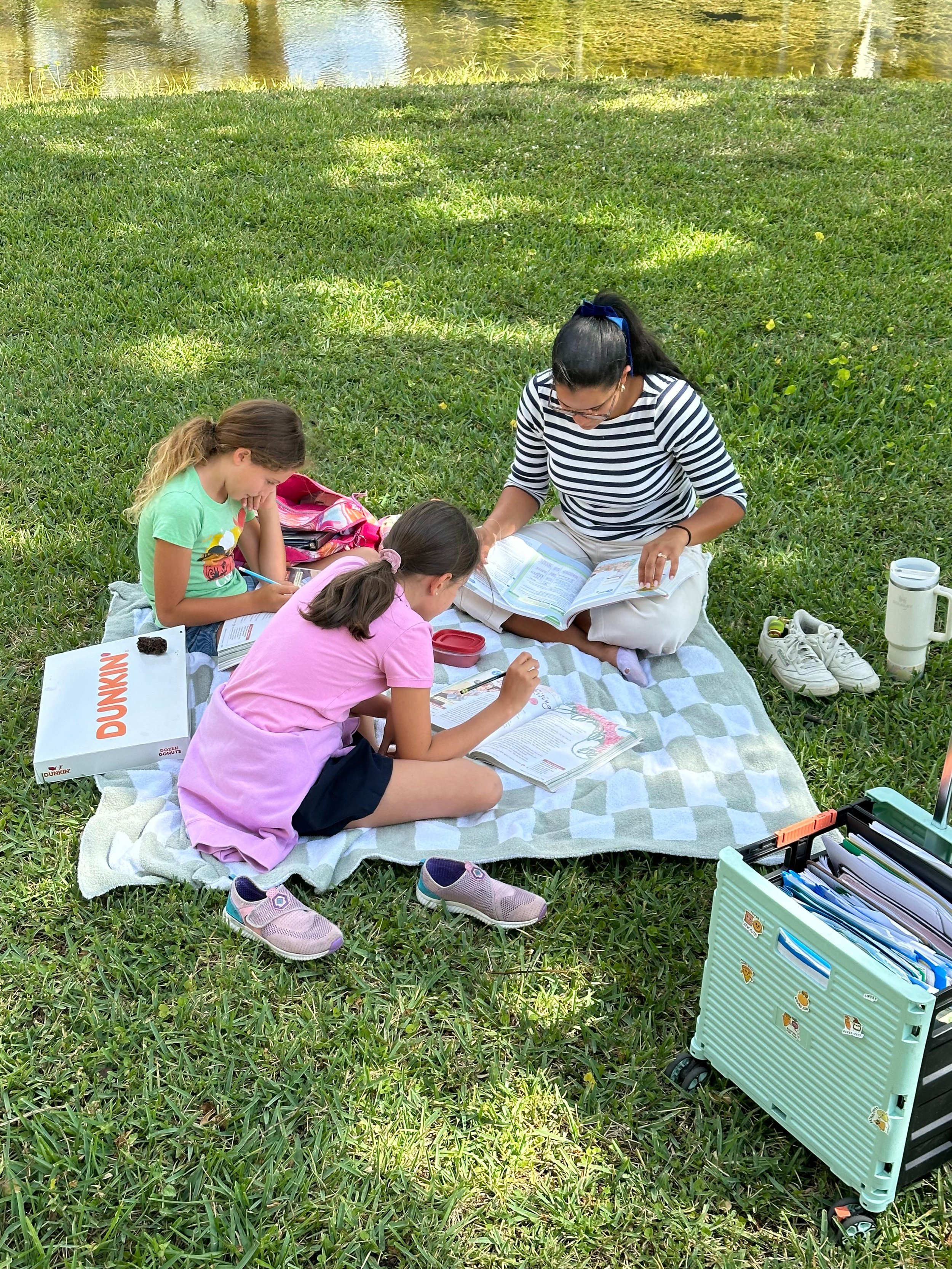 A woman and two young girls are sitting on a blanket outdoors by a pond, reading books and doing schoolwork.