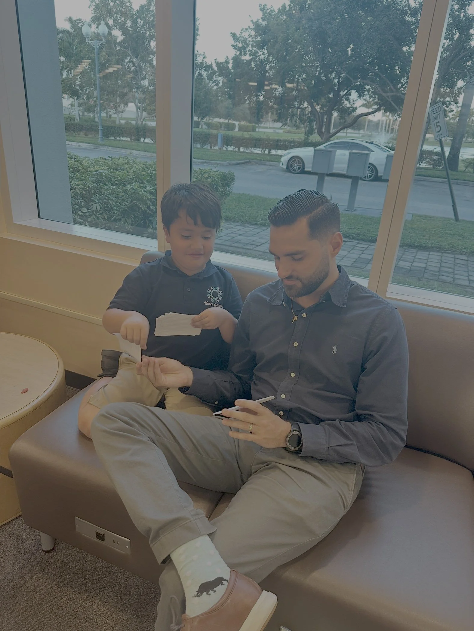 A man and a young boy sitting on a bench inside a building near large windows, looking at a phone and a piece of paper. Outside the window, there are trees, a parked white car, and a street with a speed limit sign.