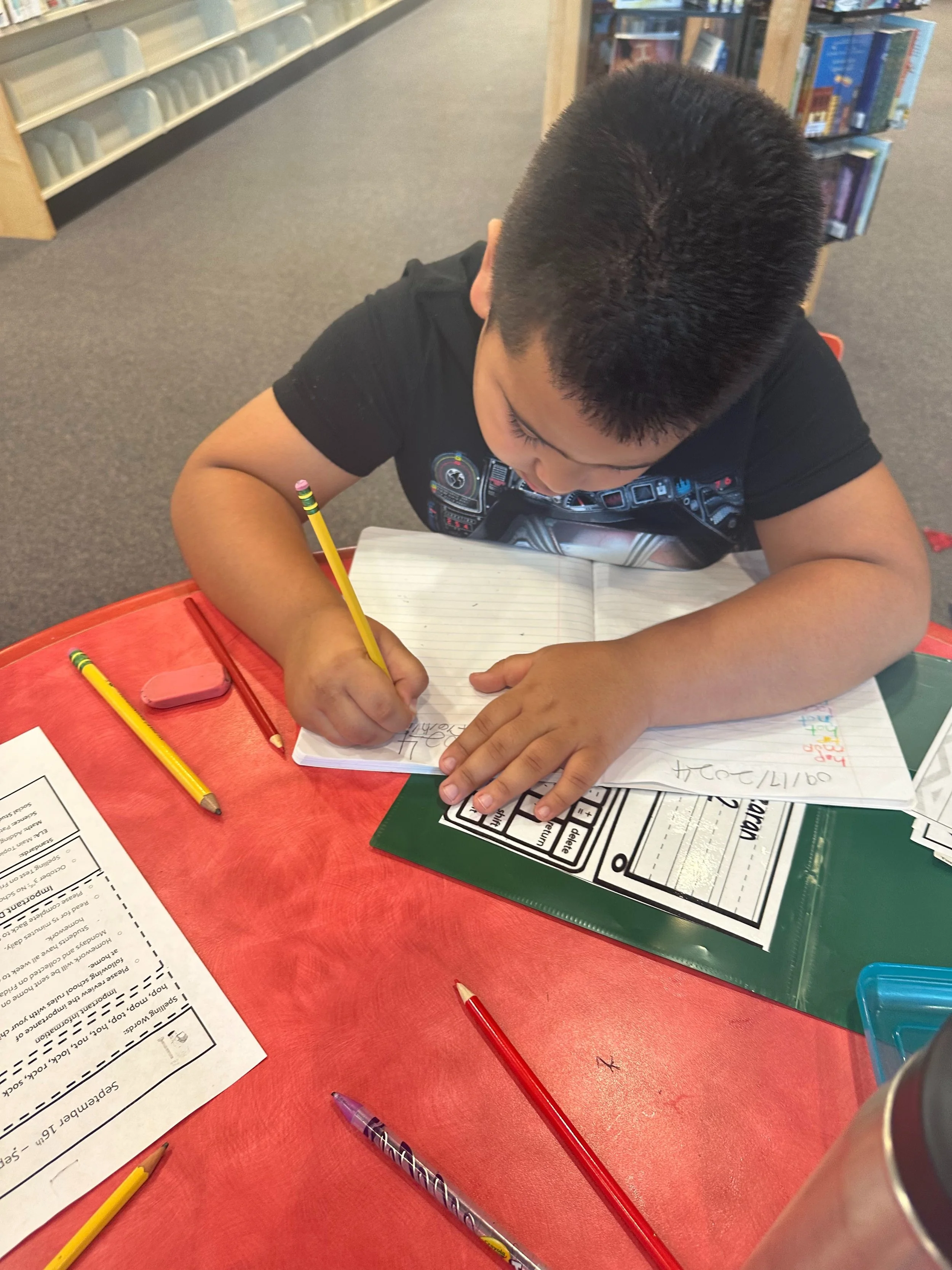 A young boy sitting at a red table in a library, writing in a notebook with a yellow pencil. There are other pencils, an eraser, and worksheets on the table.