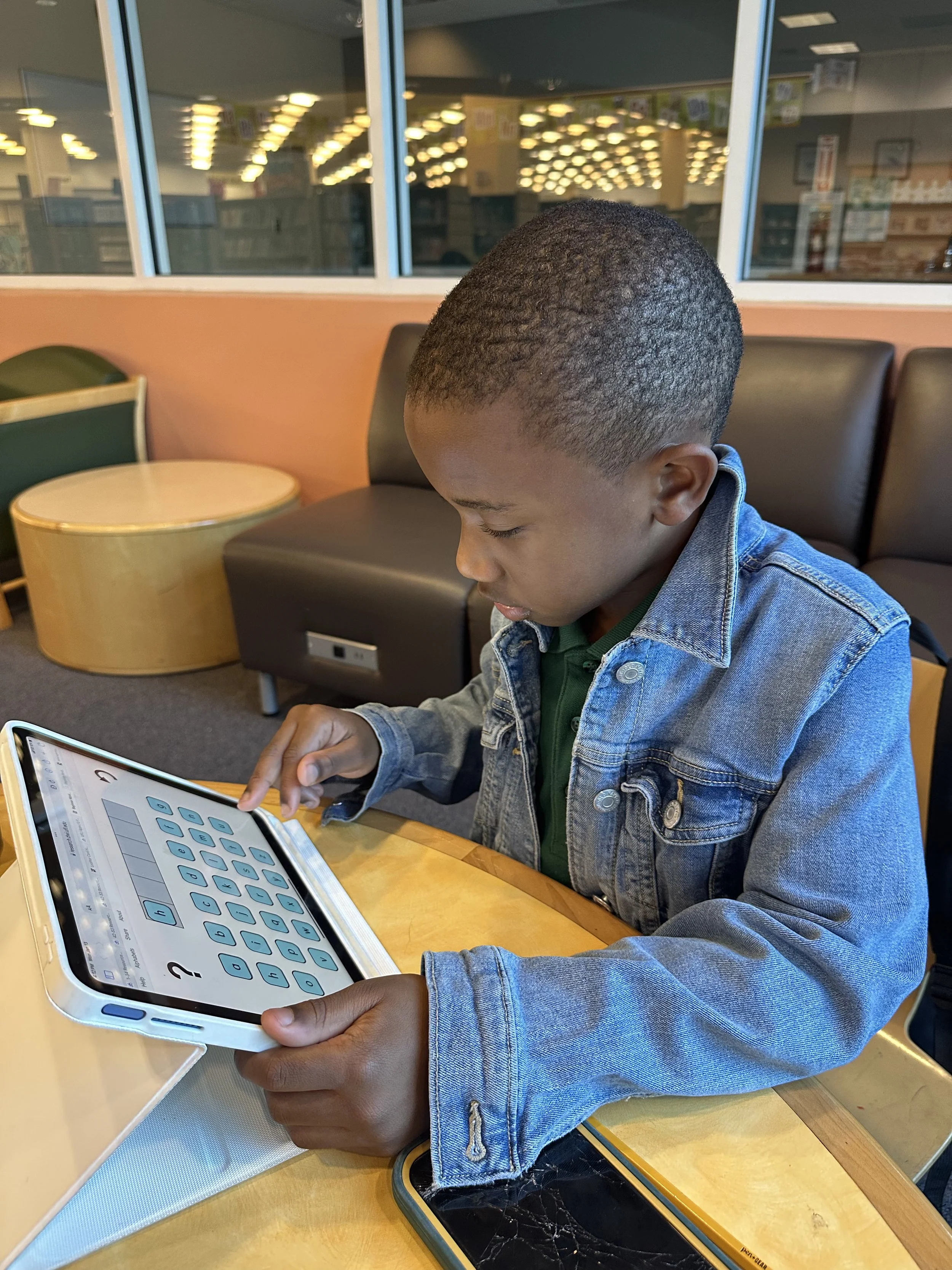 A young boy sitting at a table in a fast food restaurant or cafe, using a tablet with an educational app, wearing a denim jacket and green shirt.