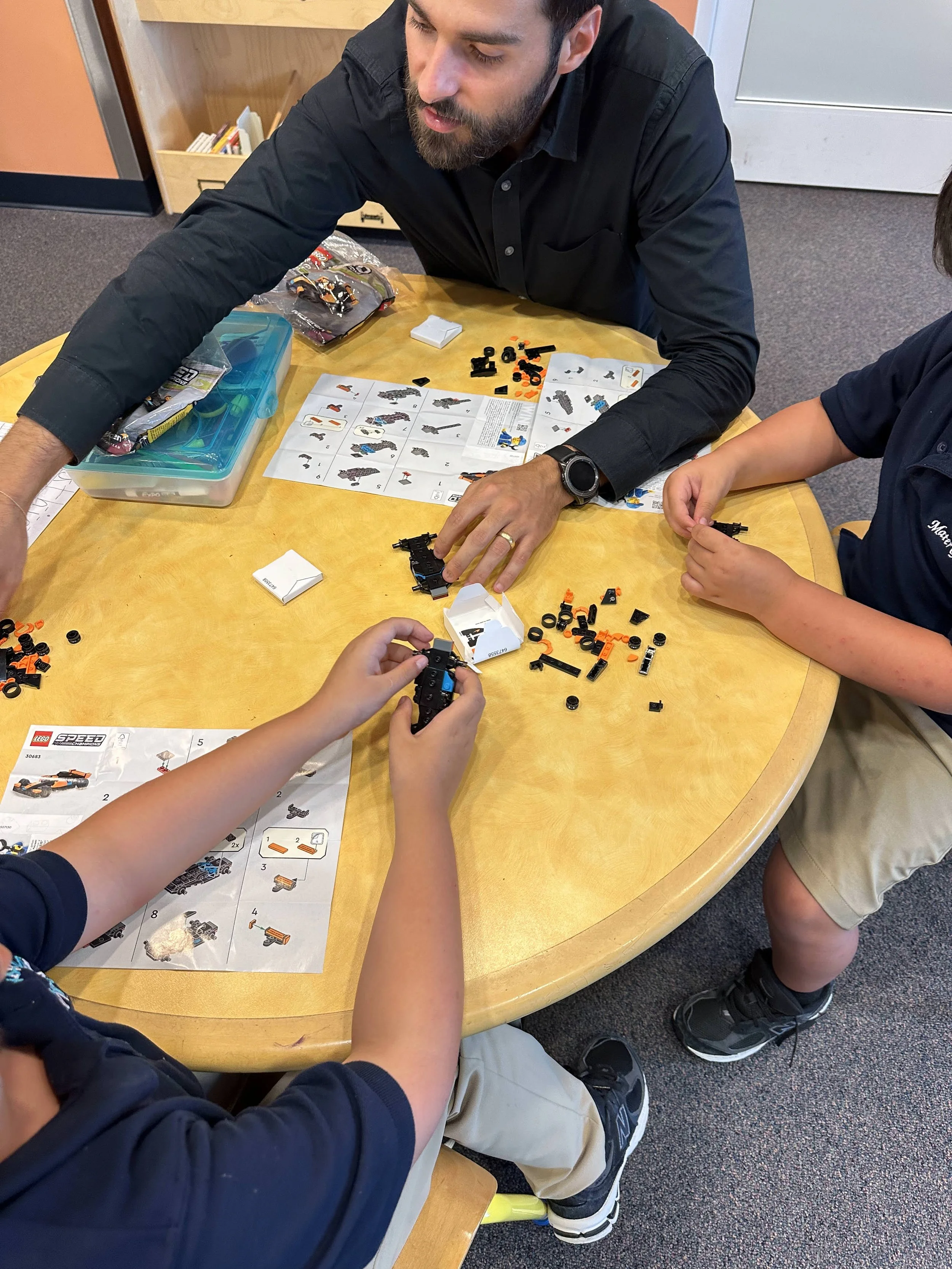 Two children and an adult assembling a Lego set at a round table with instruction sheets and Lego pieces.