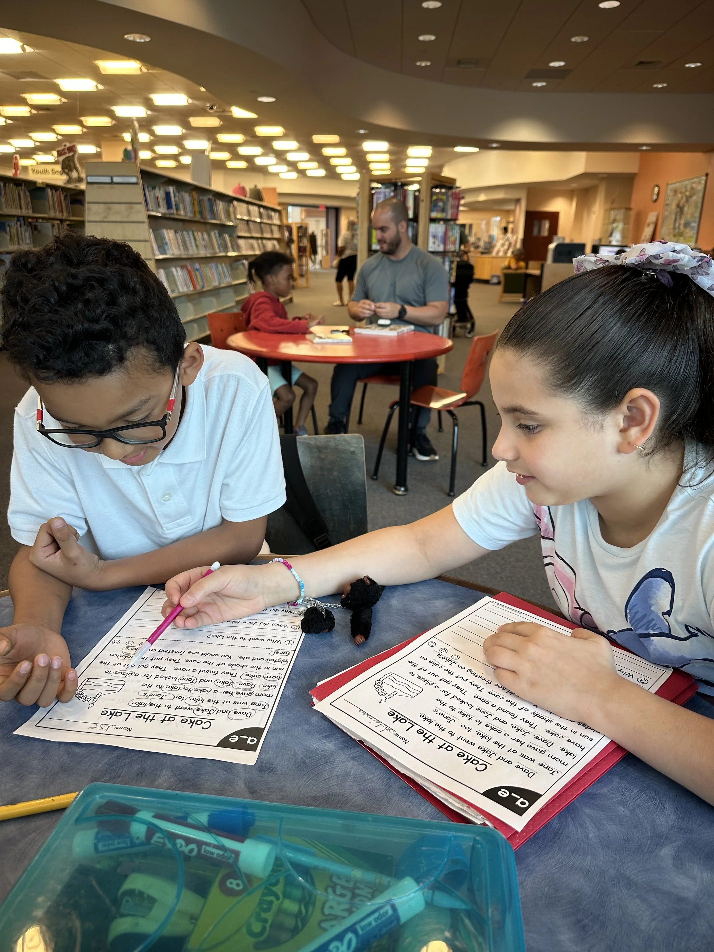 Children working on an educational activity at a table in a library. Two children in the foreground are engaged with worksheets, with bookshelves and other people in the background.