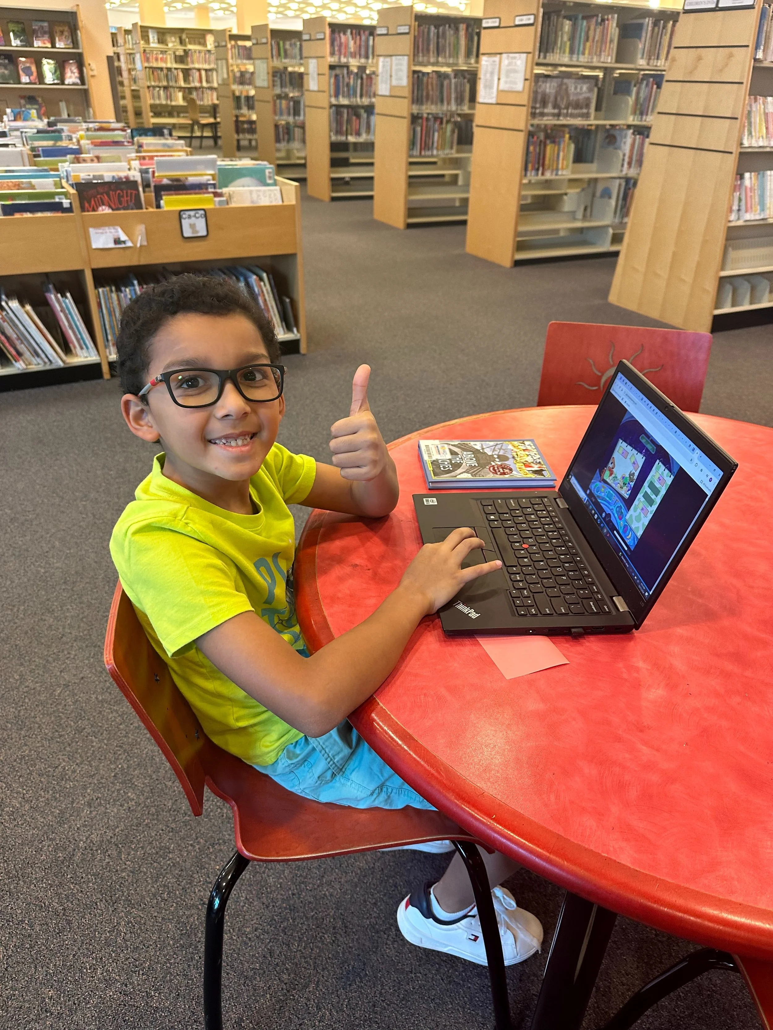 A young boy with glasses sitting at a round red table in a library, smiling and giving a thumbs-up while using a laptop.