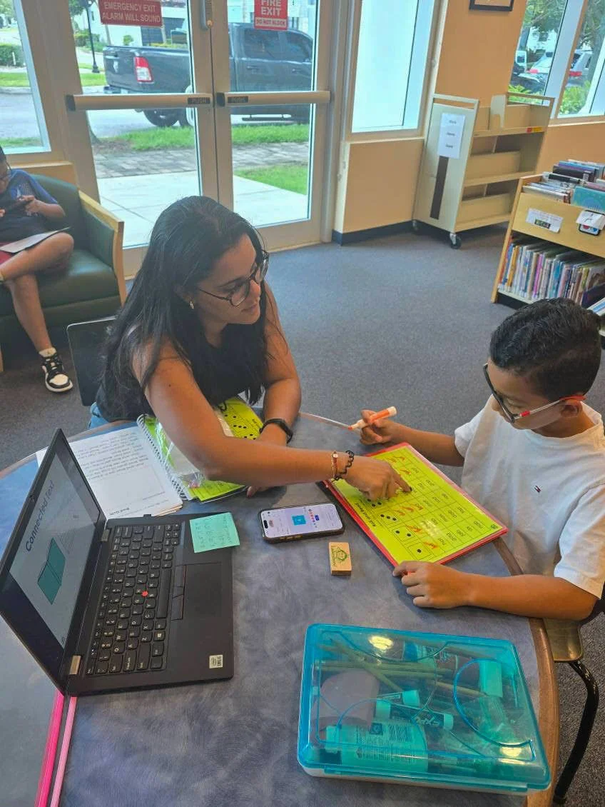 A woman and a boy sit at a table inside a library or classroom, working together on a yellow bingo game. The woman is wearing glasses and a sleeveless orange top, while the boy is also wearing glasses and a white shirt. A laptop, a smartphone, and an open book are on the table, and a blue plastic container with supplies is in the foreground. In the background, another child is sitting on a green couch, and large windows and bookshelves are visible.