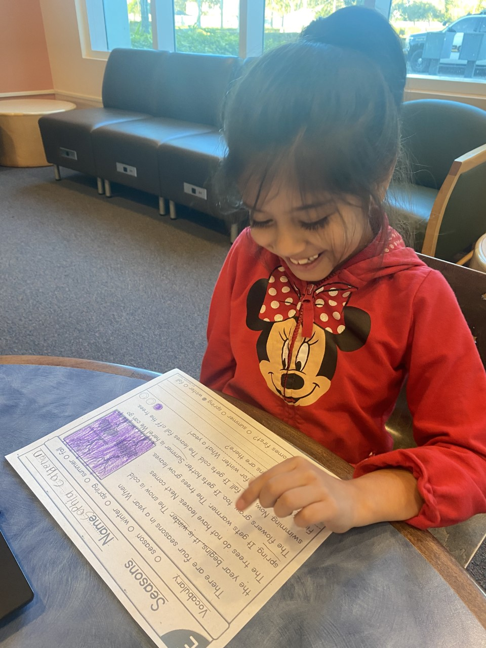 A young girl with dark hair tied back, smiling, sitting at a table and pointing to a worksheet with a purple region, in a room with large windows, a black sofa, and a parking lot visible outside.