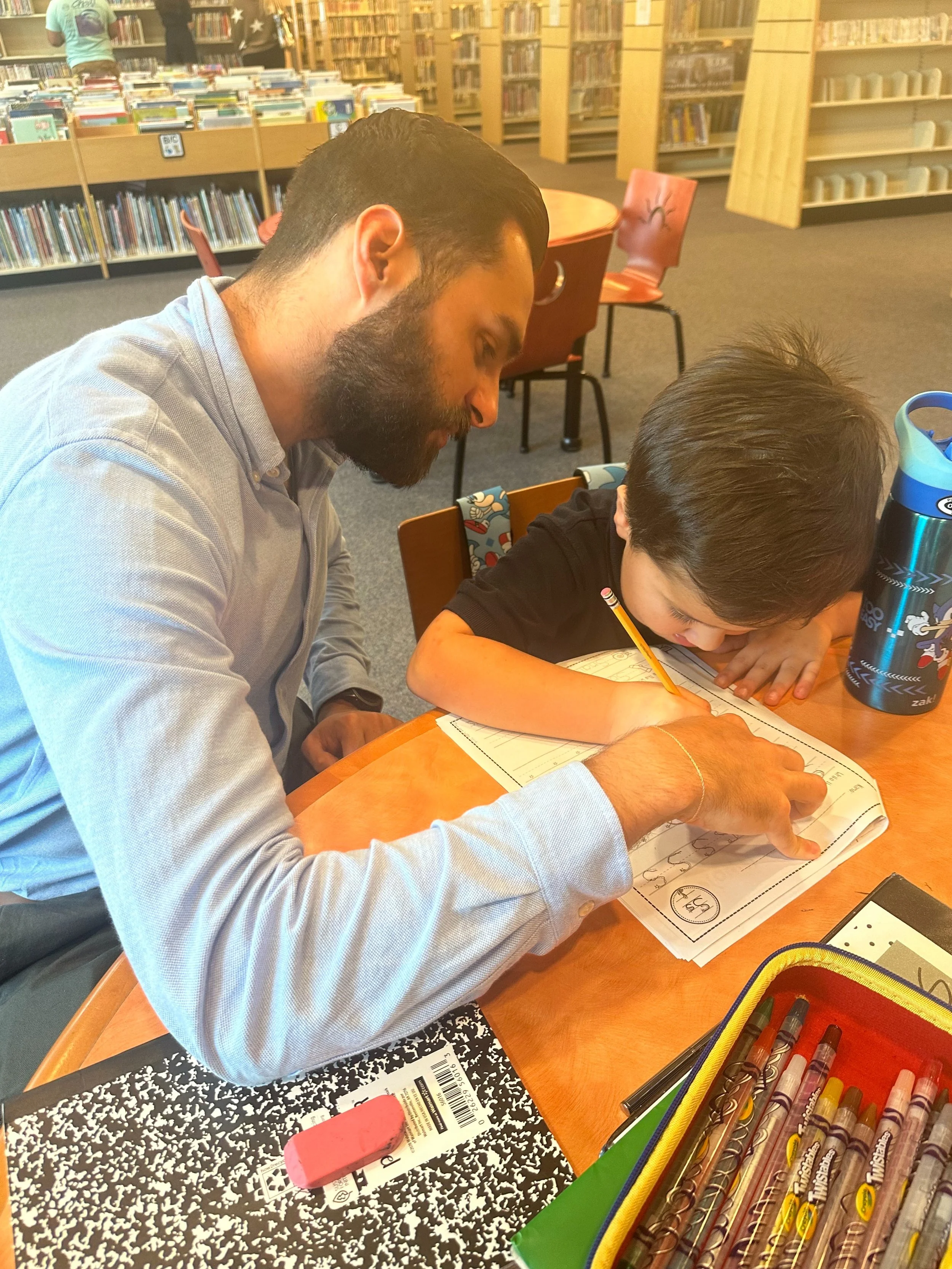 A man and a young boy sitting at a table in a library, working together on homework or a drawing. The man is guiding the boy who is writing with a pencil, and both are focused on the paper.