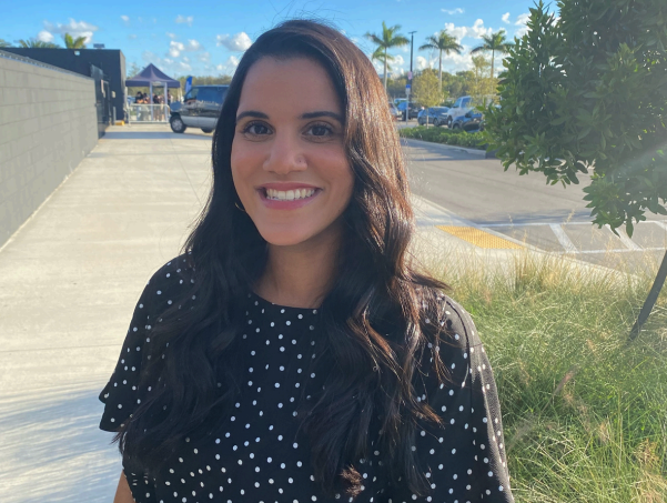 A smiling woman with long dark hair, wearing a black dress with white polka dots, standing outdoors on a sunny day near a sidewalk and green bushes.