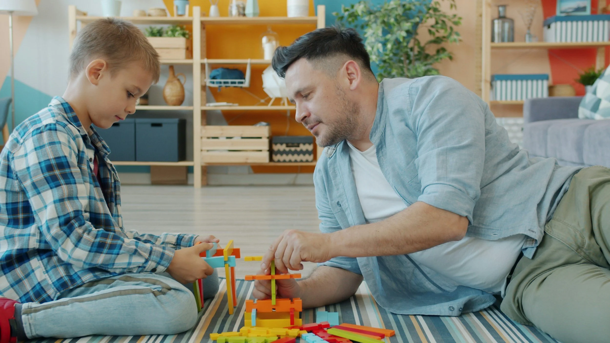 A father and son playing with colorful building blocks on the floor in a bright, modern living room.