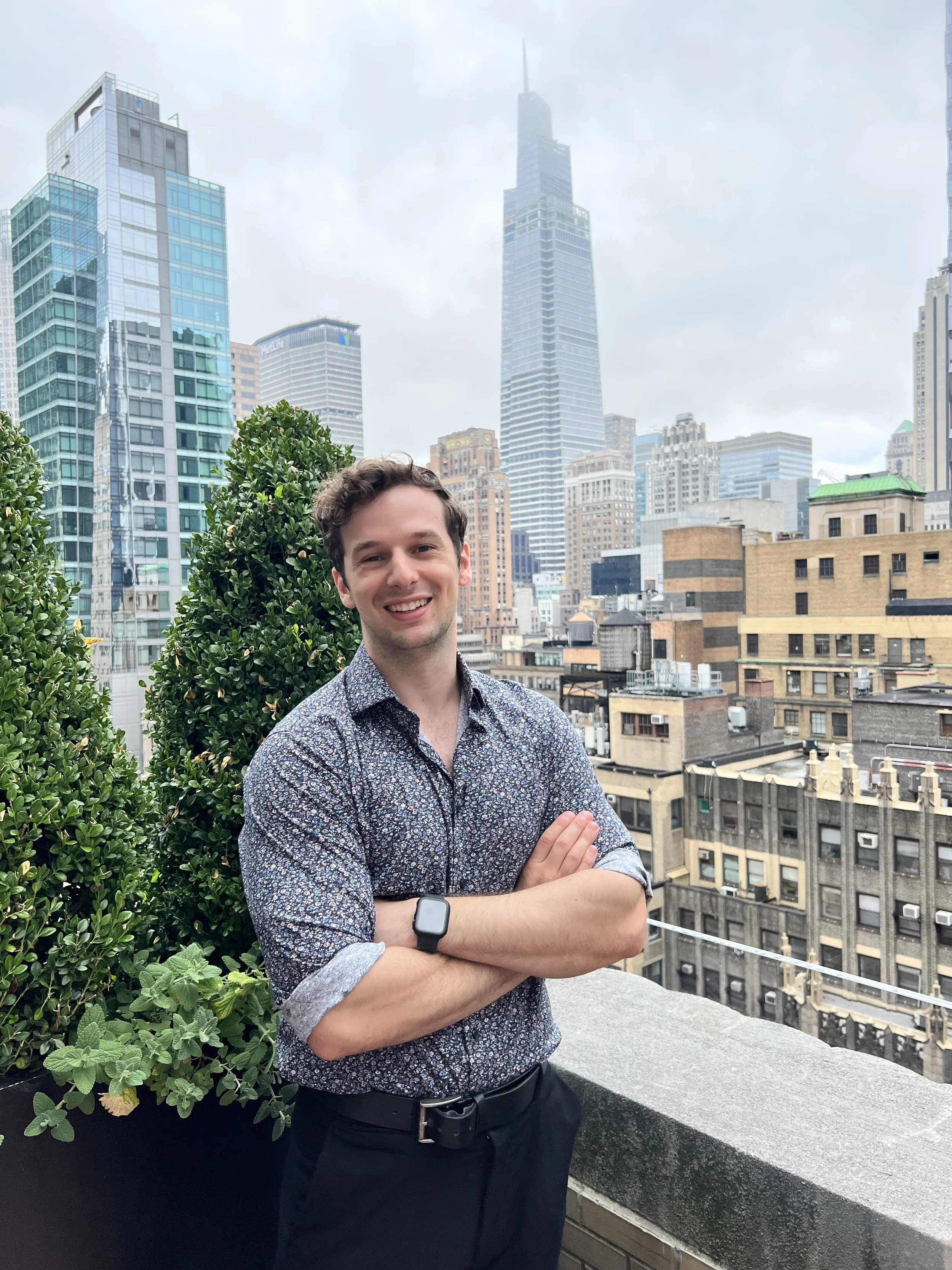 A smiling man with dark, curly hair stands on a rooftop terrace with plants and a city skyline in the background, including the One World Trade Center in New York City.