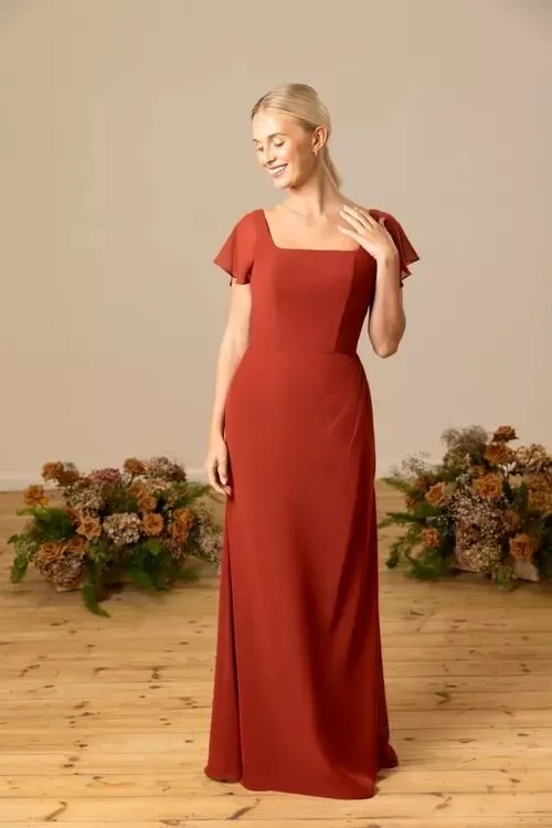 A woman with blonde hair in a low ponytail, wearing a long, rust-colored dress with short flutter sleeves, standing on a wooden floor with flower arrangements behind her, smiling and touching her shoulder.
