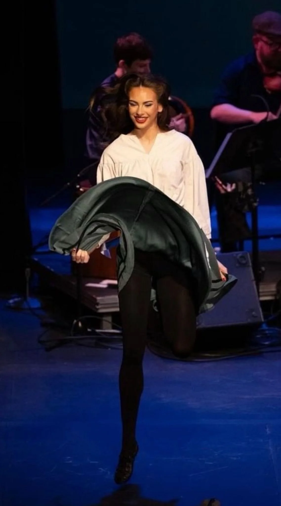 A woman on stage holding a coat, with a joyful expression and a smile. In the background, there are musicians preparing to perform.