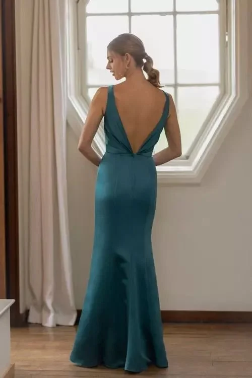 Woman in a teal backless gown standing by a window, looking to the side.
