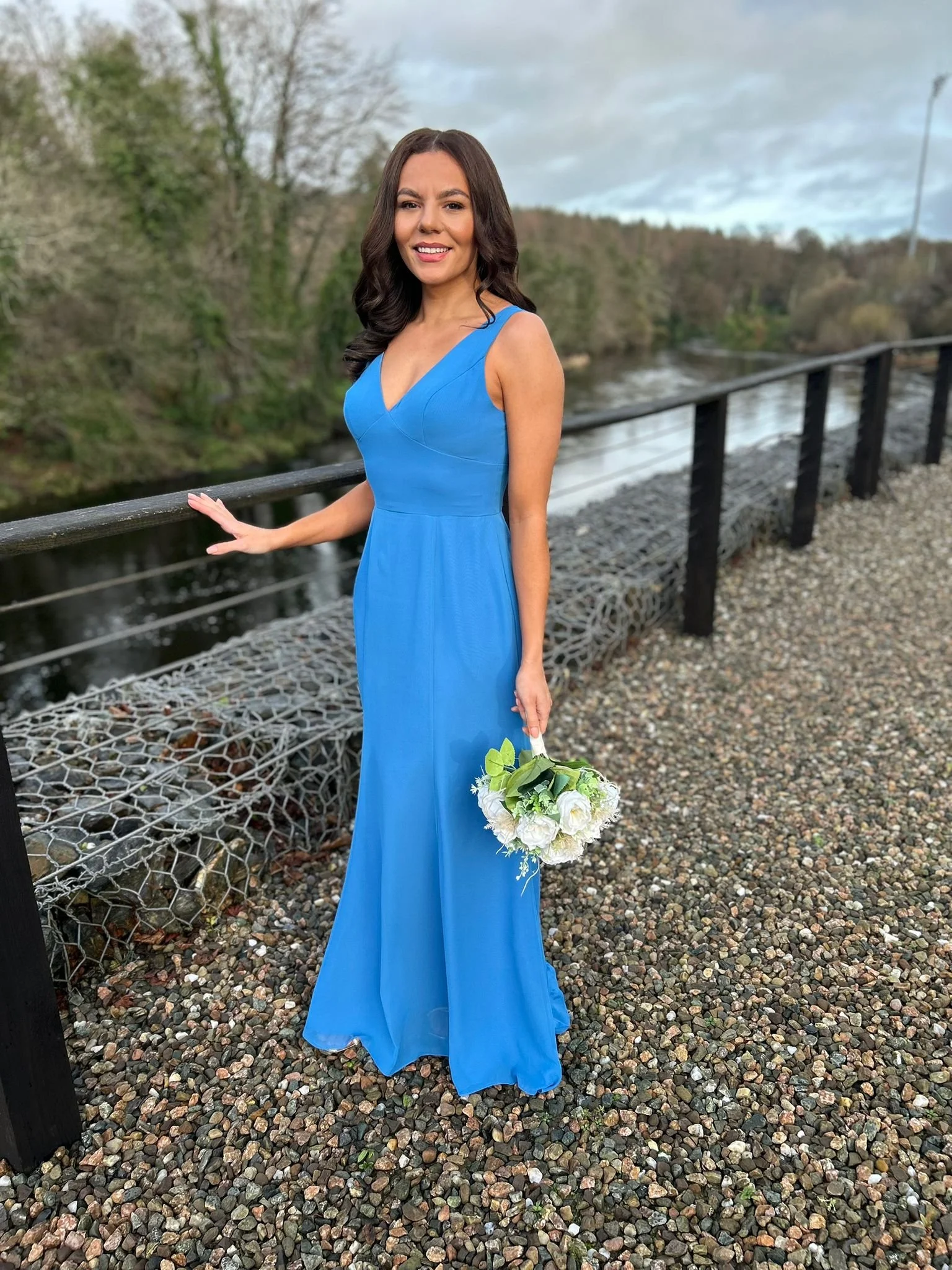 A woman in a long blue dress holding a white flower bouquet, standing on a gravel path near a railing by a river, with trees and cloudy sky in the background.