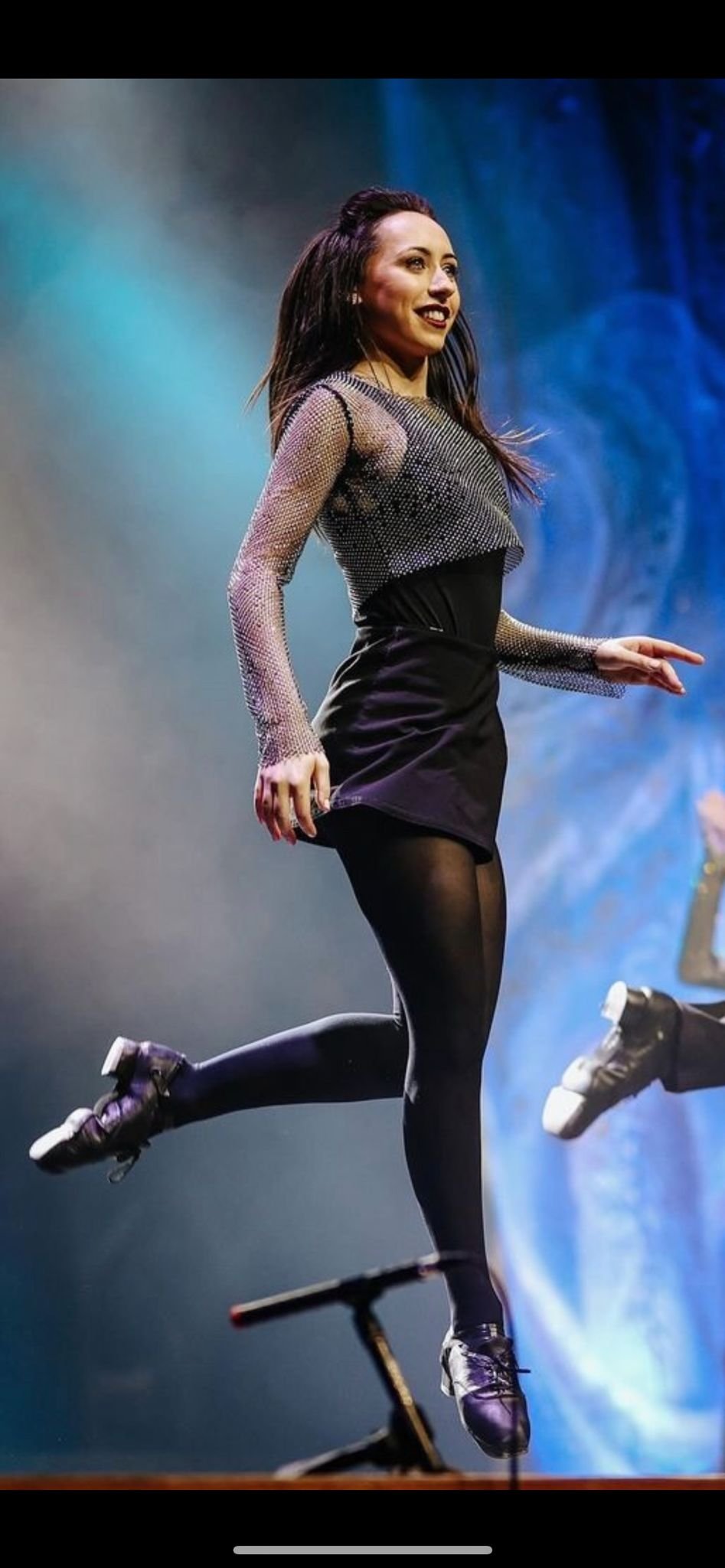 A woman skating on stage at an event, smiling and wearing a flashy, metallic long-sleeved top, black skirt, and tights, with a colorful background.