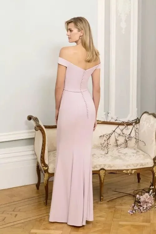 A woman in an off-shoulder pink gown with buttons down the back, standing in a room with a vintage sofa and floral decorations.