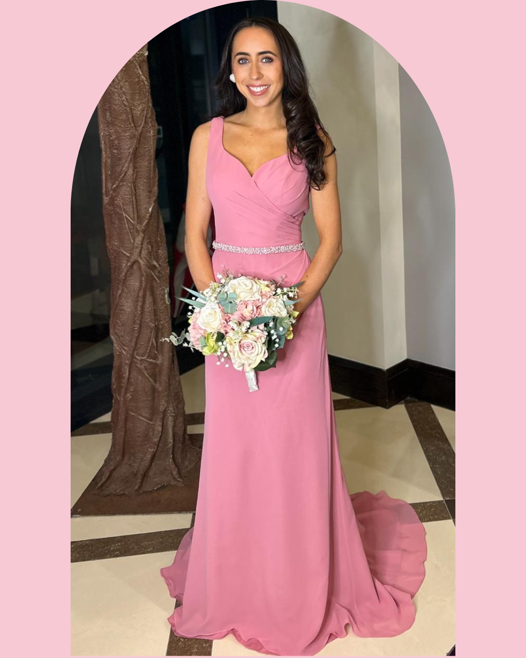 A woman in a pink evening gown holding a bouquet of white and pink roses, standing indoors with a decorative background.
