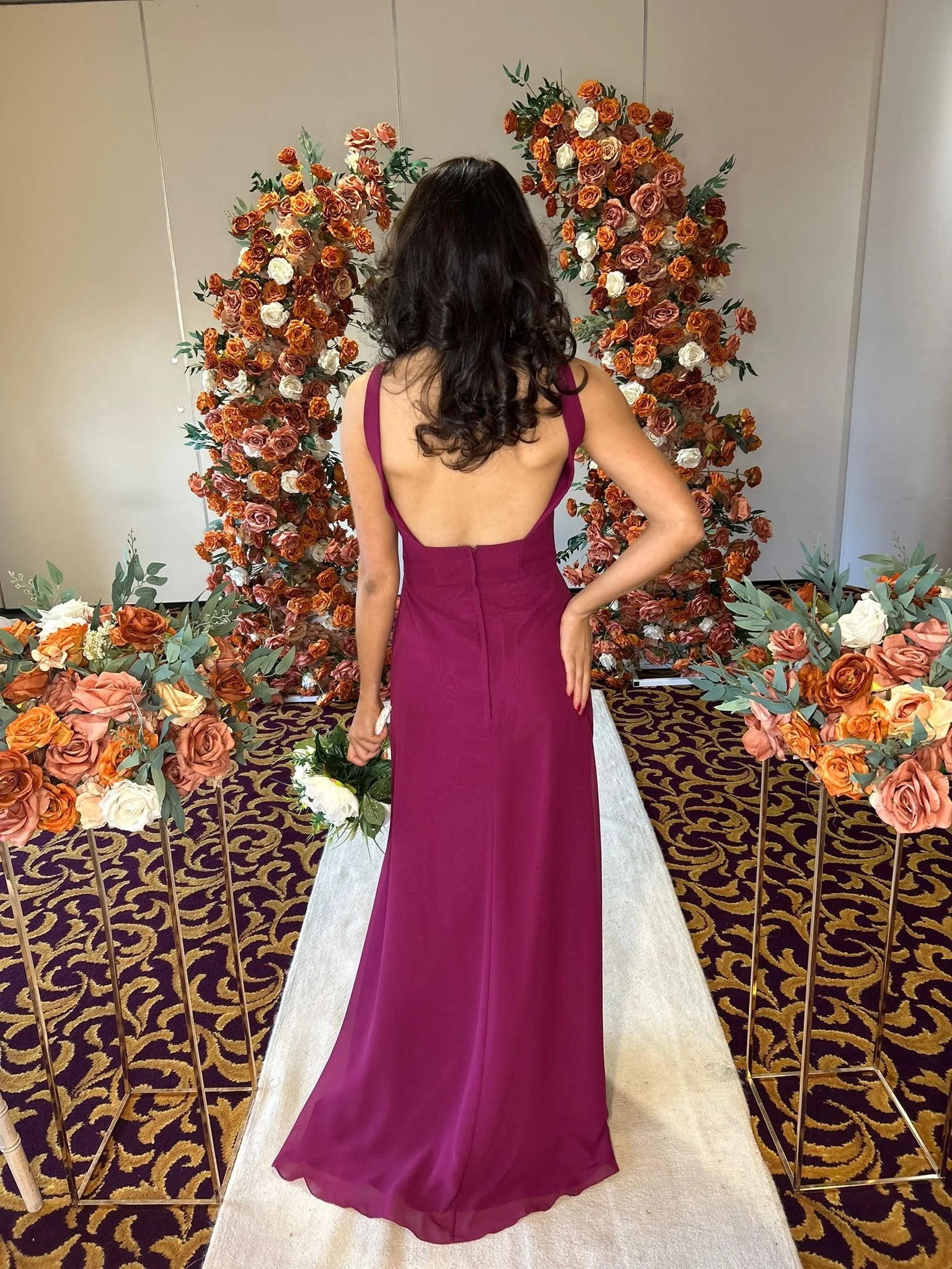 A woman in a long magenta dress is standing on a white aisle with floral arrangements on either side, and a floral arch behind her.
