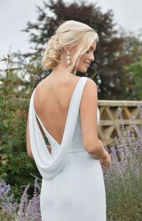 A woman with blonde hair styled in an elegant updo, wearing a white dress with a low back and draped fabric, standing outdoors near purple flowers and greenery.