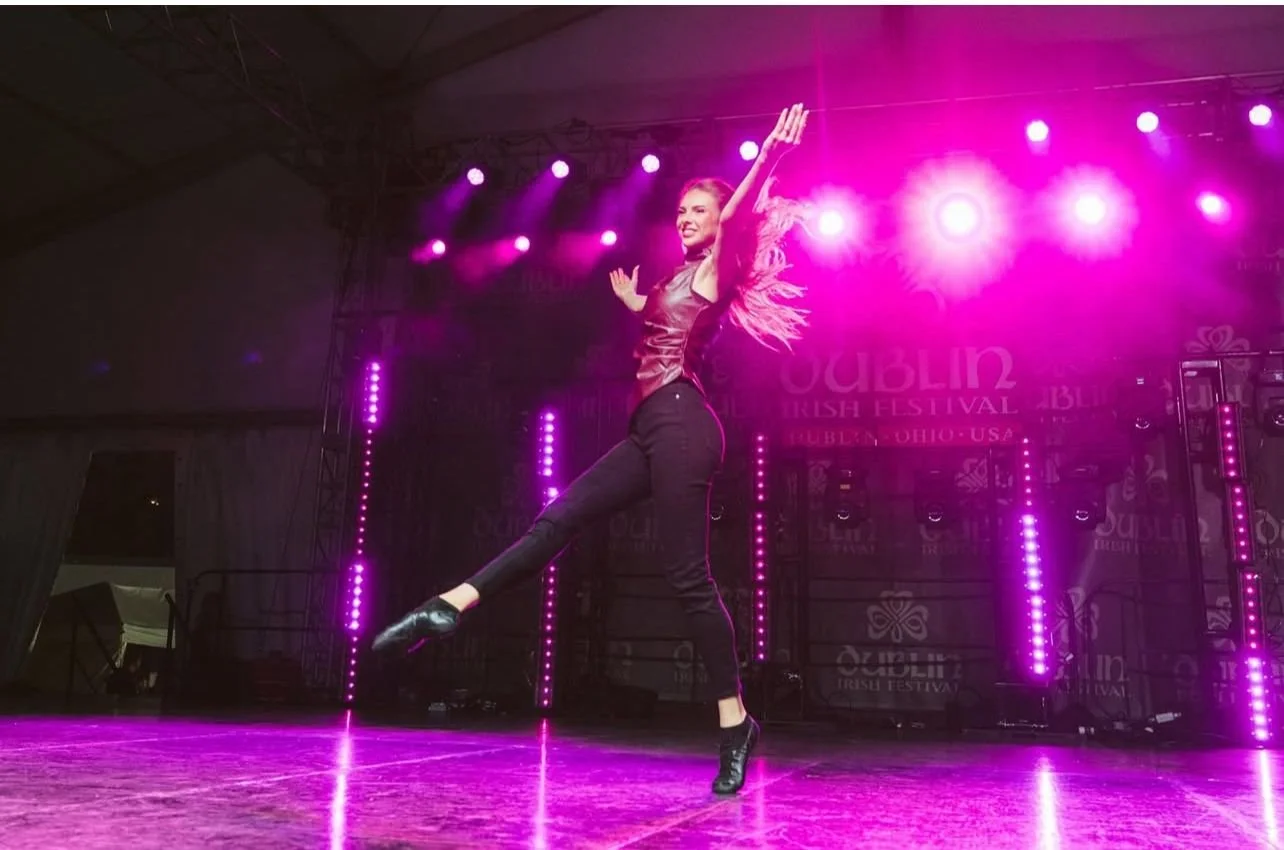 A female dancer performing on stage at an Irish festival. She is wearing dark clothing and black shoes, captured mid-dance with one leg extended and arms raised. The stage is lit with bright pink and purple lights.
