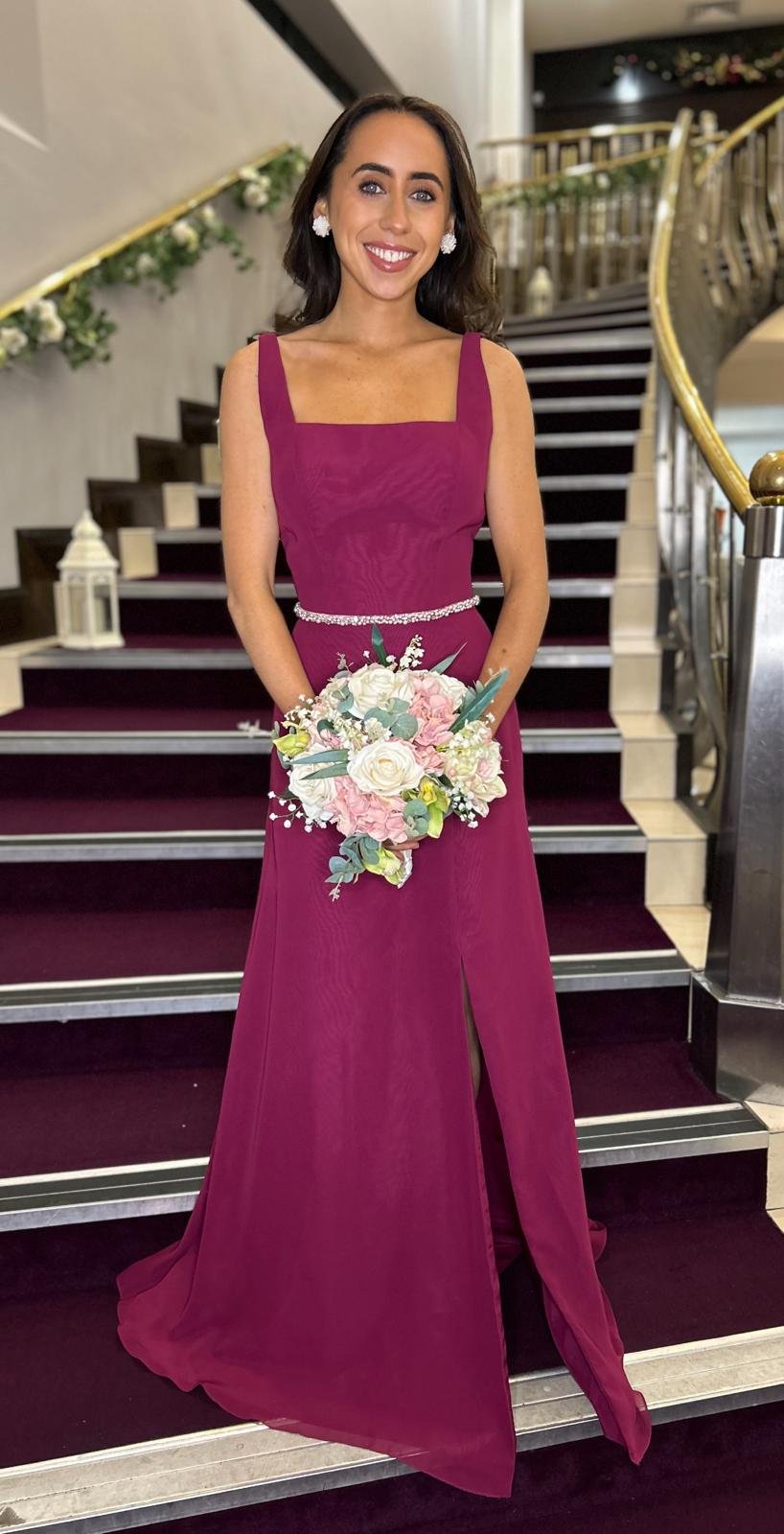 A woman in a magenta evening gown holding a bouquet of white, pink, and green flowers standing on a staircase decorated with white flowers and lanterns.