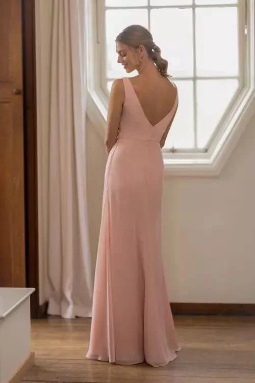 A woman in a pink gown standing indoors near a window with curtains, with her back to the camera, looking down.