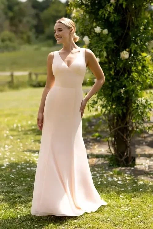 A woman in a white sleeveless dress standing outdoors on grass with trees and a fence in the background.